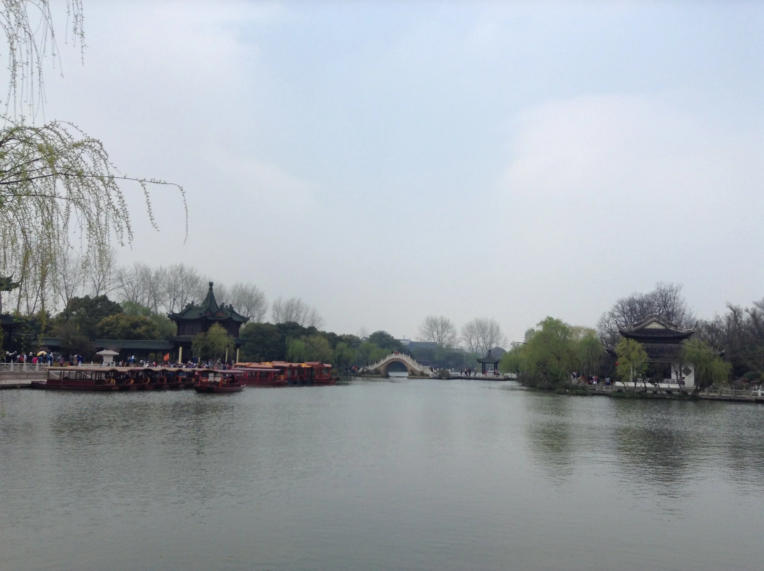 A serene lake scene with traditional Chinese architecture, a small bridge, and boats, surrounded by trees, under a cloudy sky.