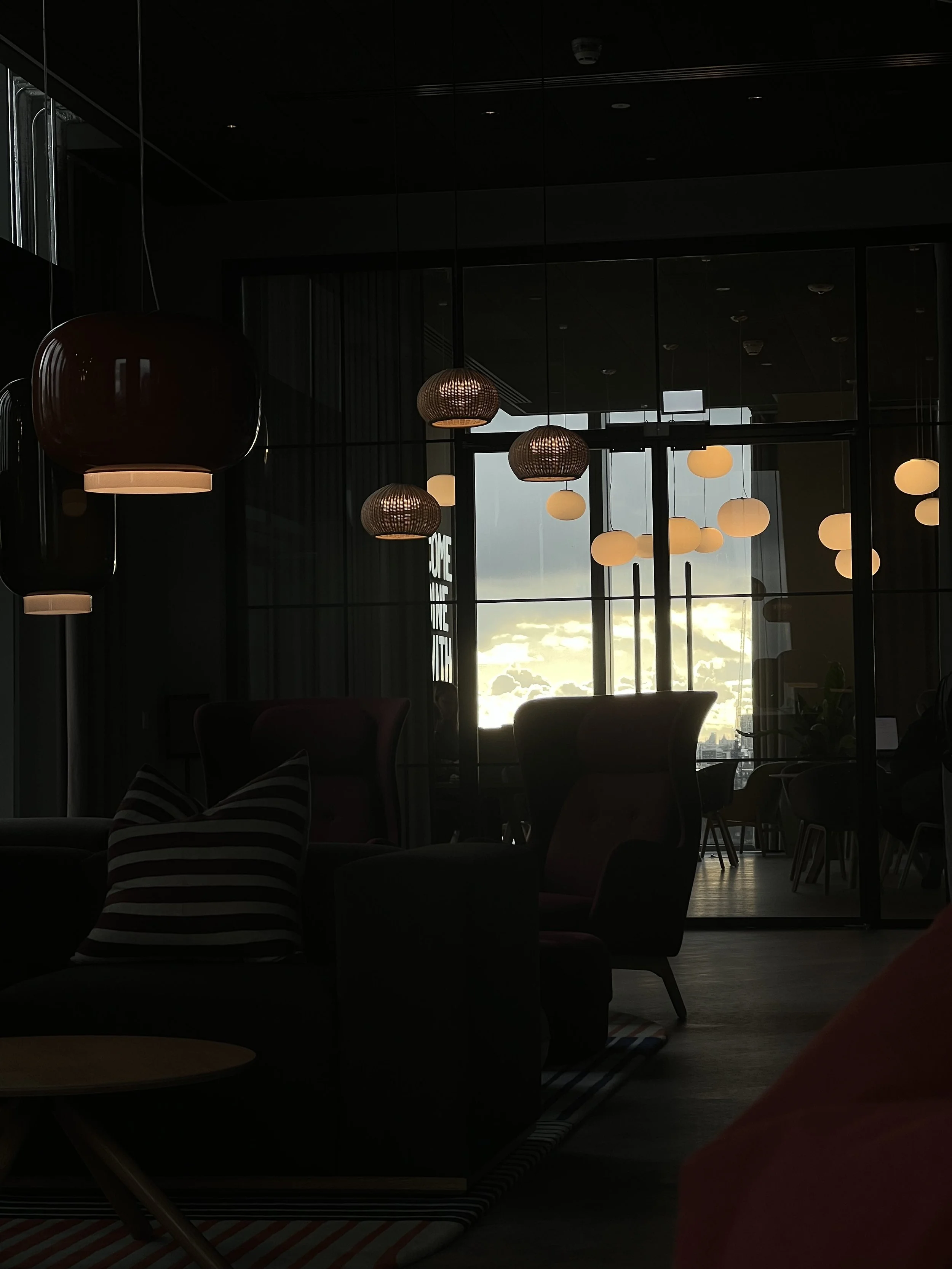 Dark interior of a modern building with hanging pendant lights and large windows showing a cloudy sky during sunset.