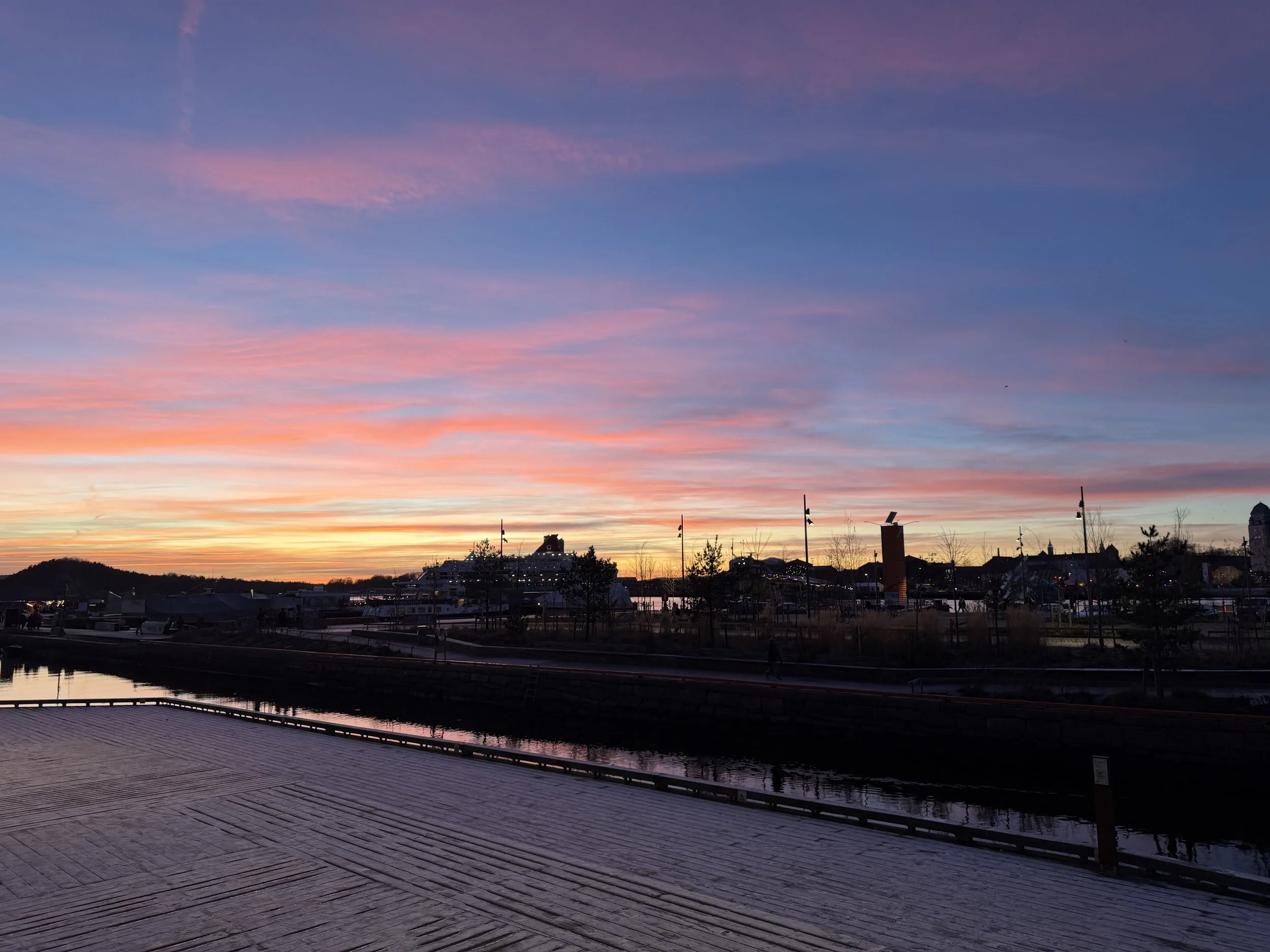 A sunset sky with pink and purple clouds over a waterfront area with boats, buildings, and trees.