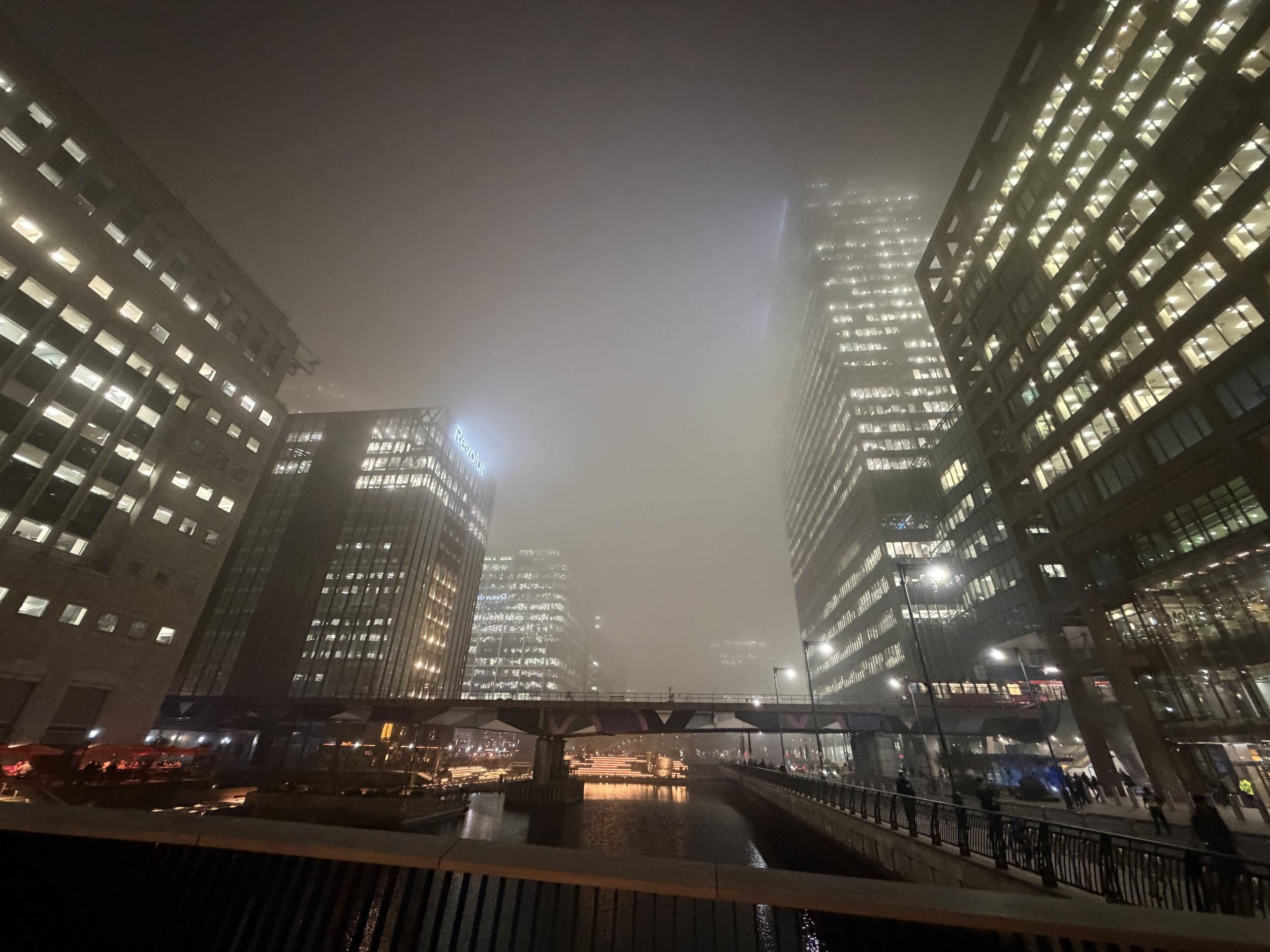 Nighttime cityscape with tall buildings, some with illuminated windows, shrouded in fog or mist, with reflections on the water below and a bridge in the foreground.