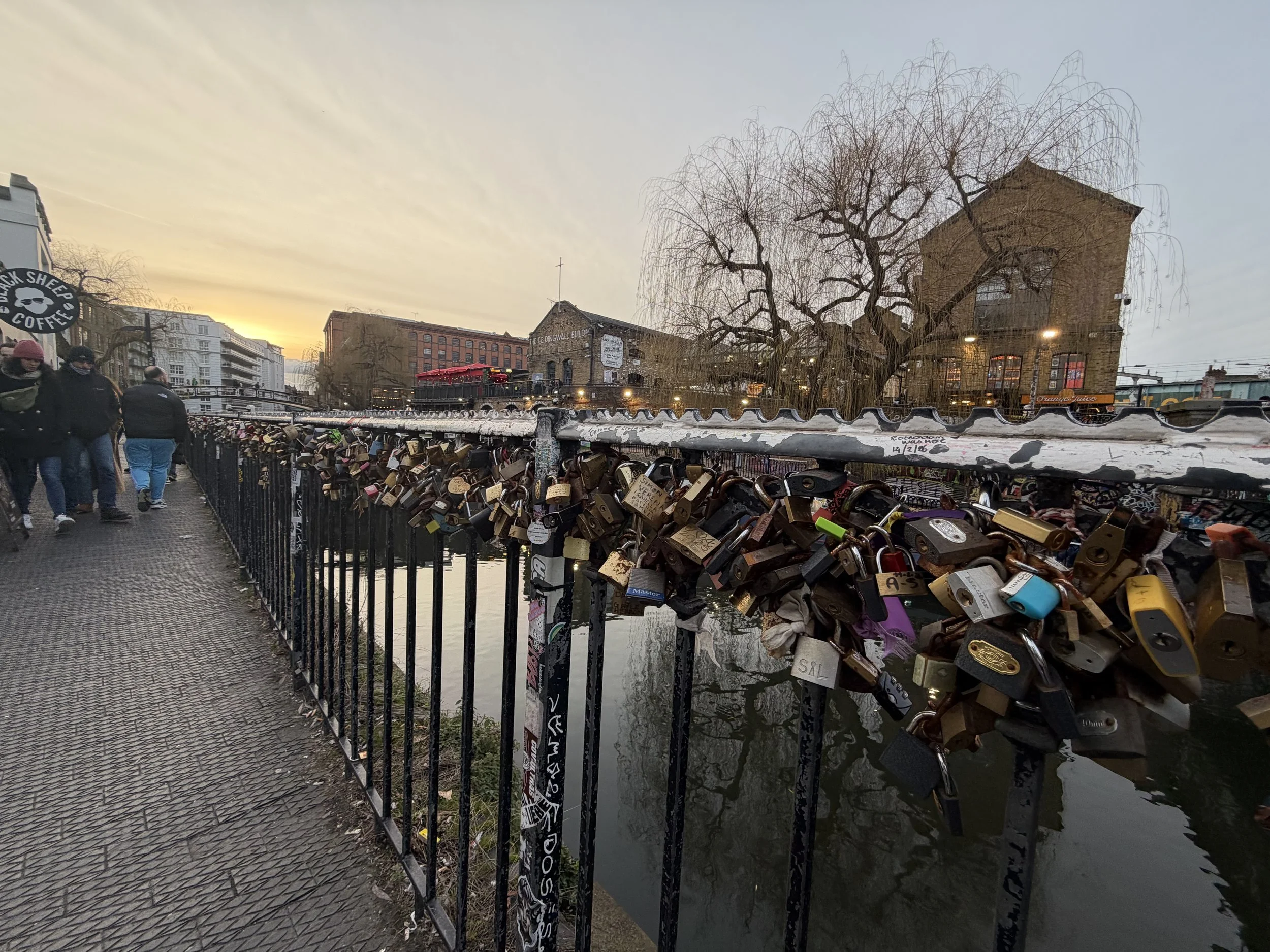 A view of a bridge over a waterway with numerous padlocks attached to the railing, some with inscriptions. On the left side, people are walking on a sidewalk. In the background, there are buildings, including a brick one with a sign reading 'The Engi