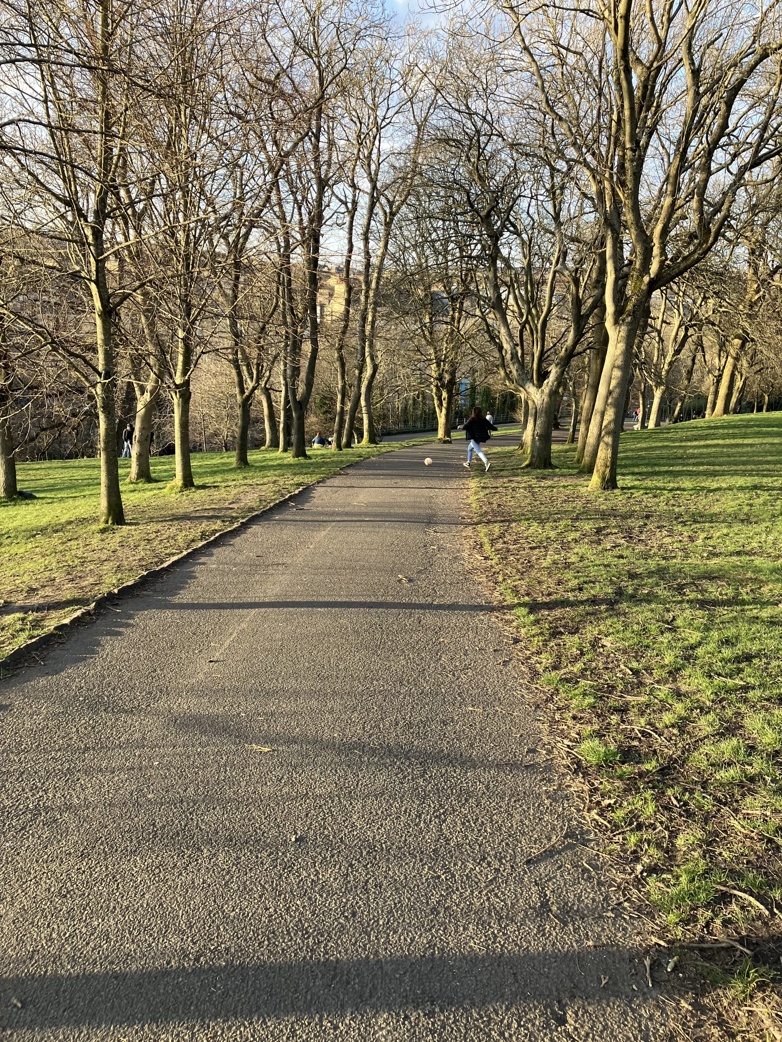 A paved pathway through a park lined with leafless trees on both sides. A girl is kicking a soccer ball on the path. Other people are visible in the background, and the sky is clear and blue.