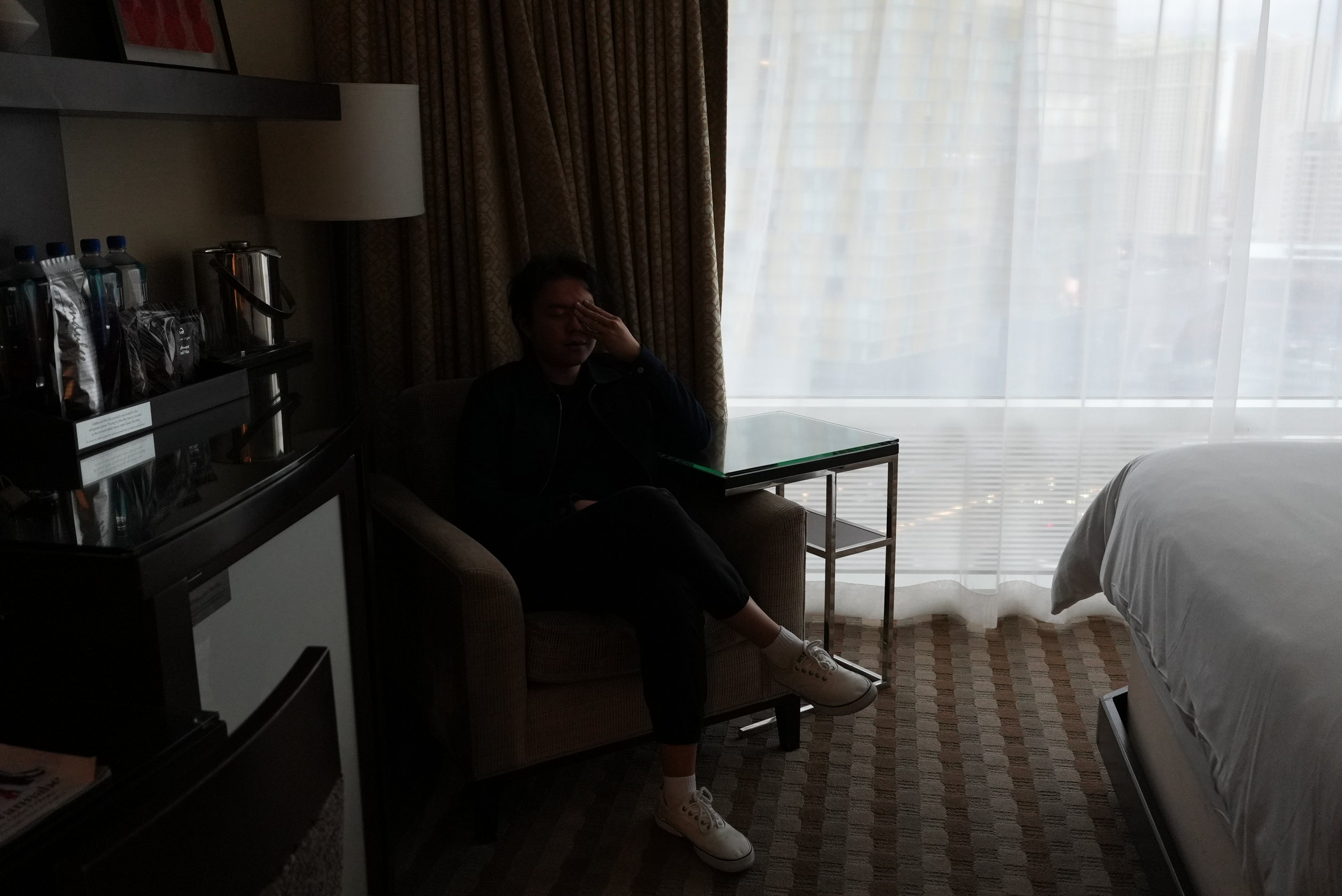 A person sitting in a hotel room near a large window with sheer curtains, covering their face with one hand, wearing white shoes and dark clothing. The room has a side table, a lamp, and a beverage station with bottled water and snacks.