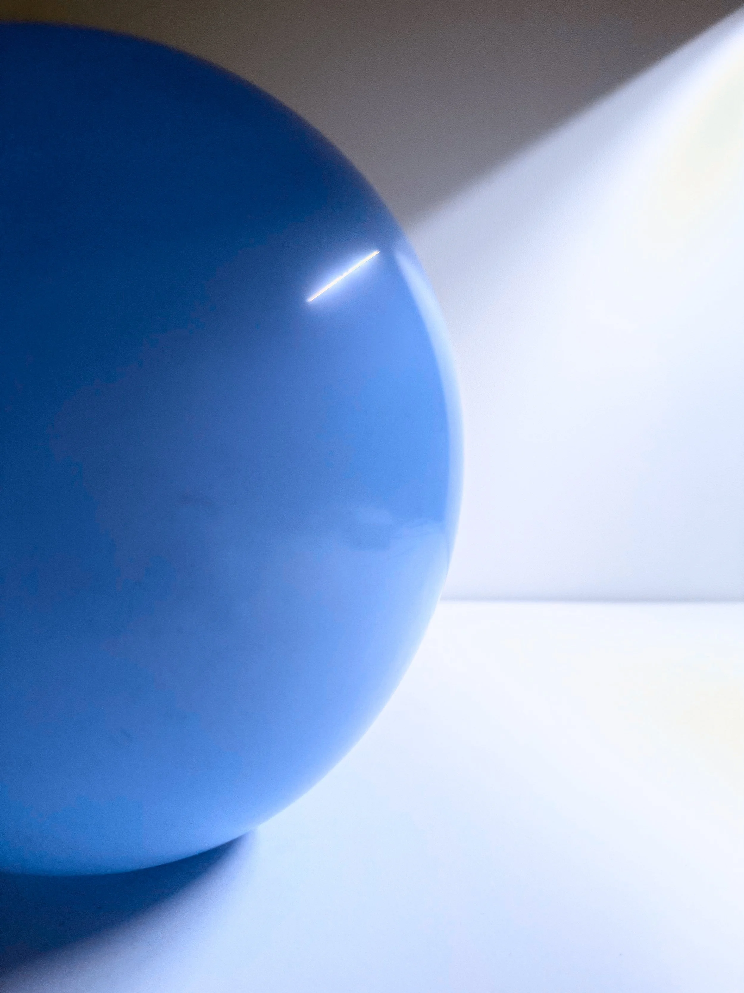 Close-up of a blue object, possibly a balloon or sphere, with a small bright reflection on its surface, against a light background.