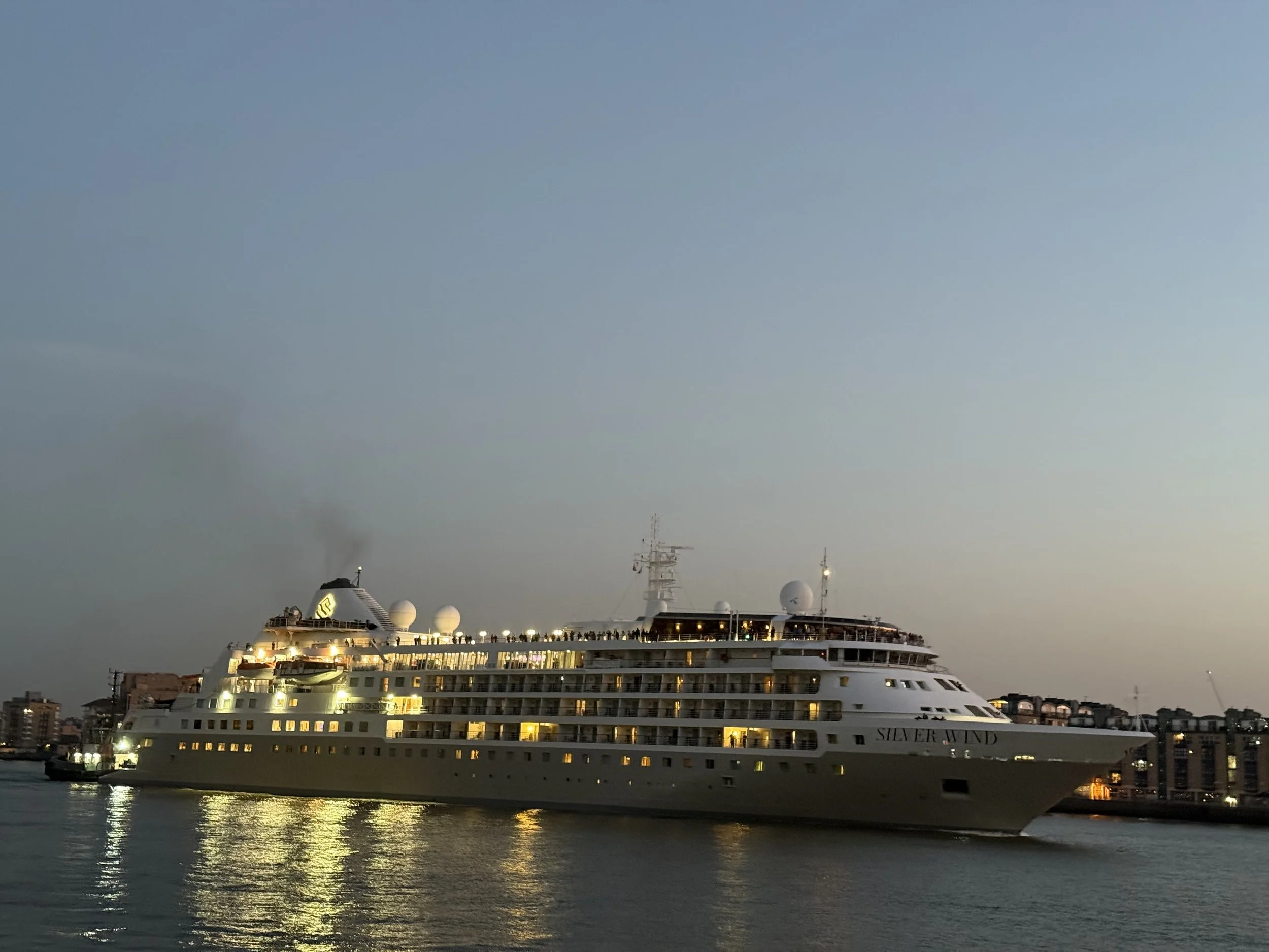A large white cruise ship named "Silver Wind" is docked at a harbor during dusk, with city buildings in the background and city lights reflecting on the water.
