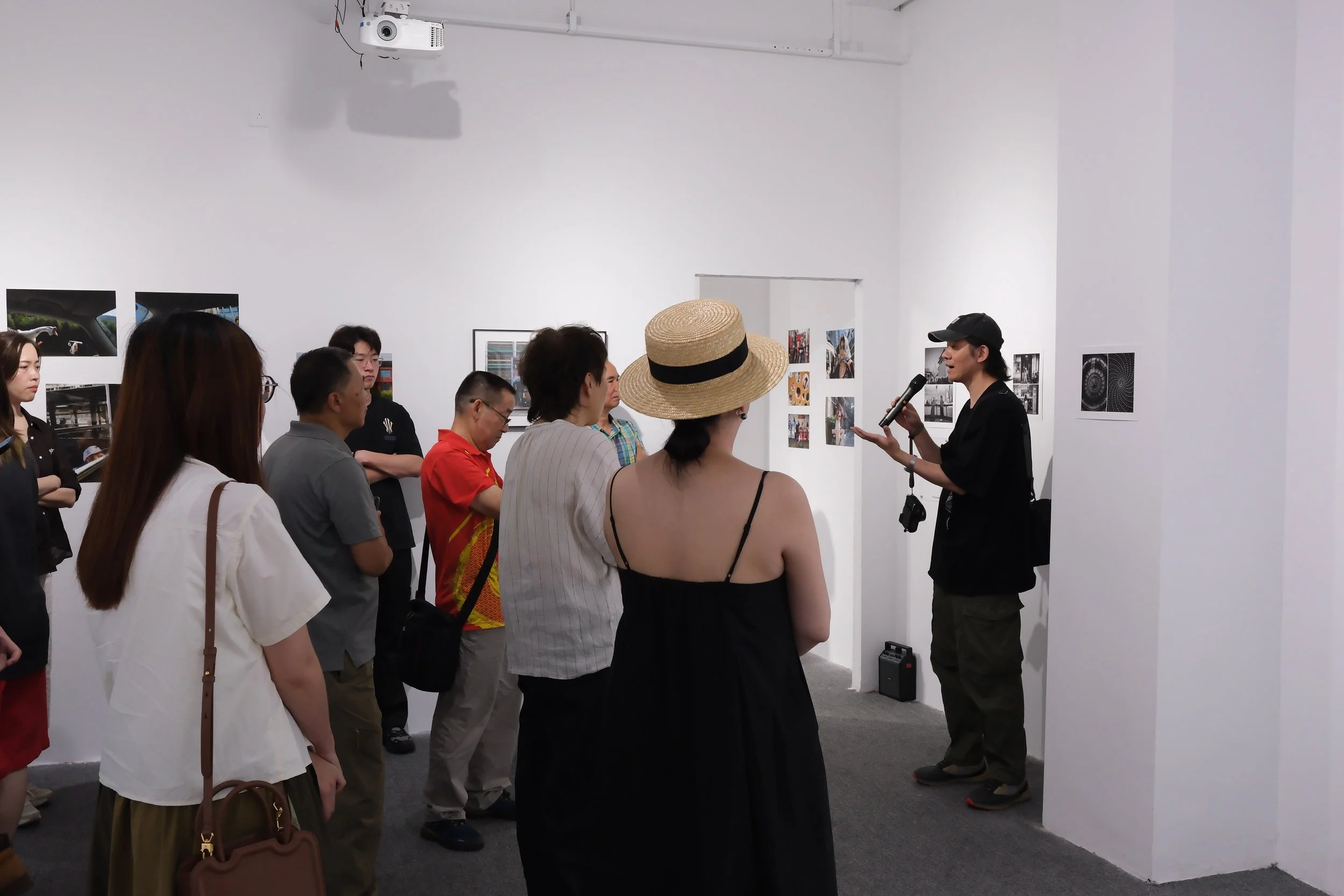 A group of people attending an art exhibition, listening to a speaker with a microphone in front of a white wall displaying various photographs and artwork.