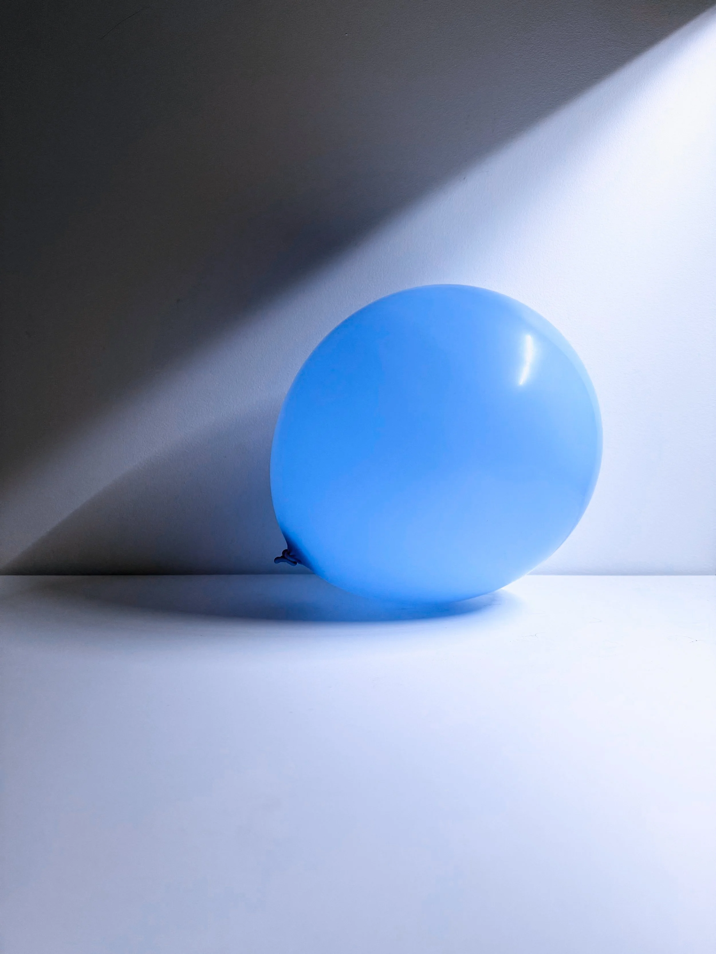 A single blue balloon on a flat surface, casting a shadow in a well-lit room with a plain background.