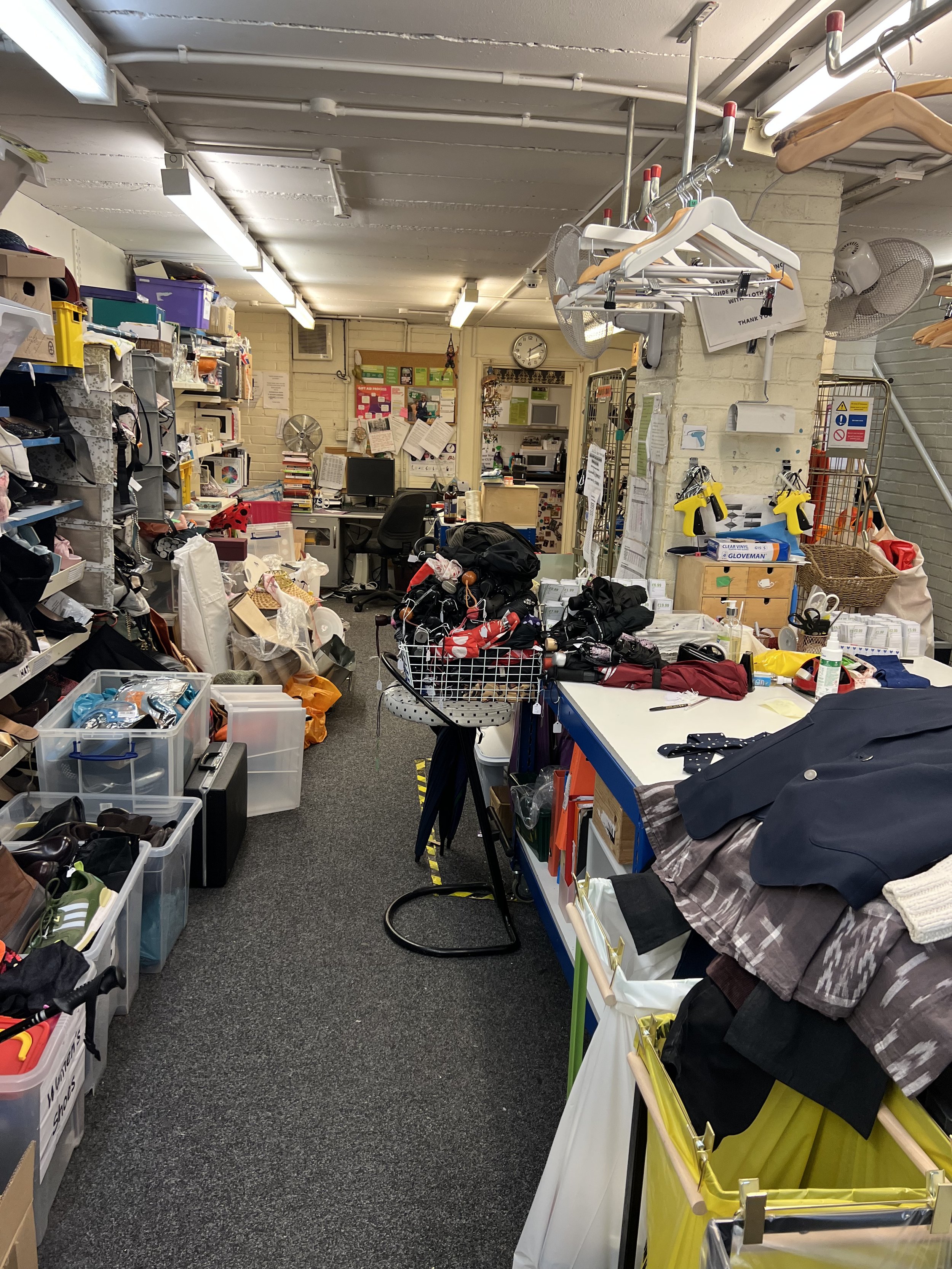 Cluttered thrift store or donation center with shelves filled with boxes and items, tables with clothes and accessories, and various signs and descriptions on the wall.