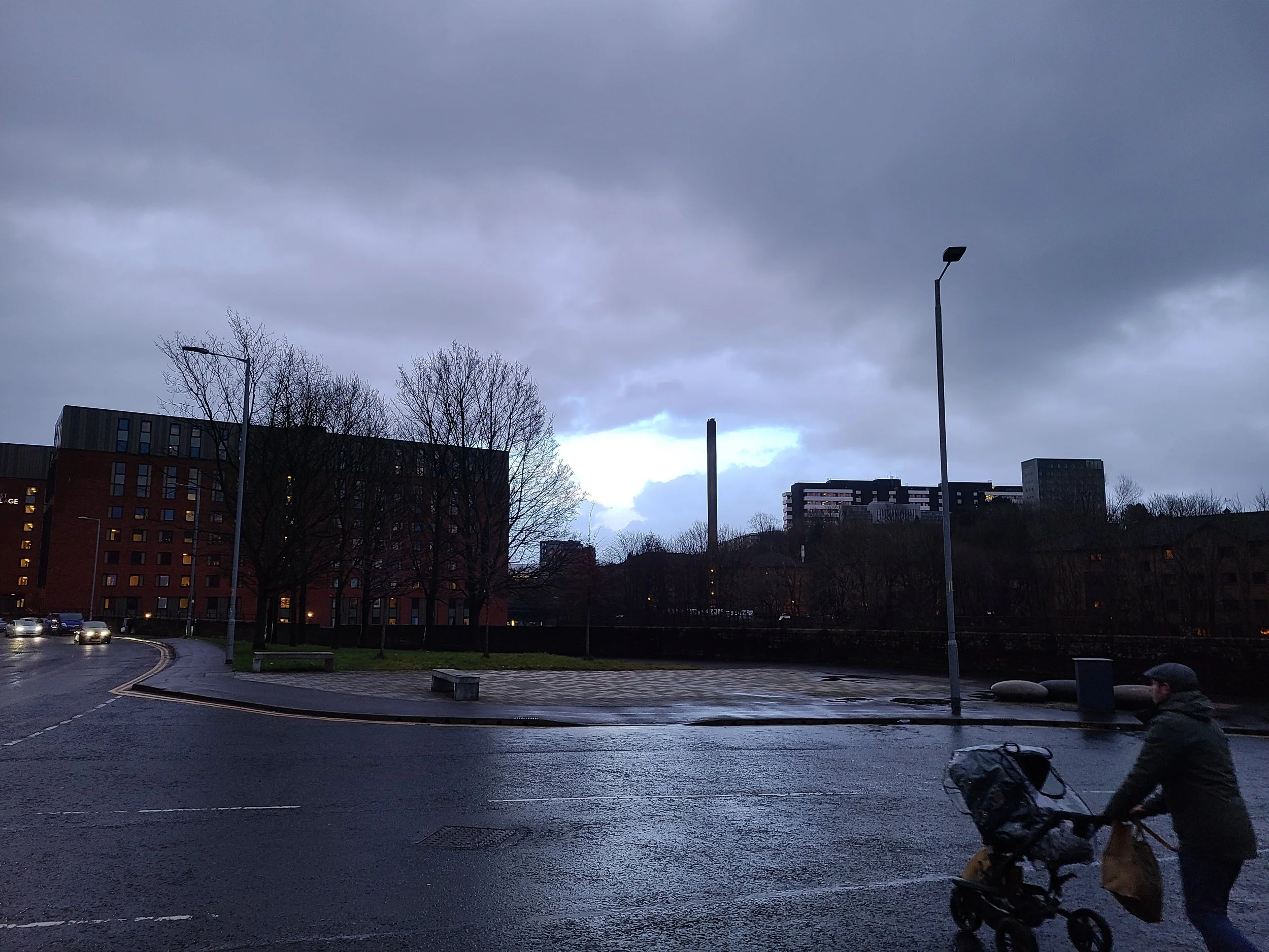A city street scene during cloudy weather, with parking lot, leafless trees, apartment buildings, street lamps, and a man sitting on the curb with a bicycle.