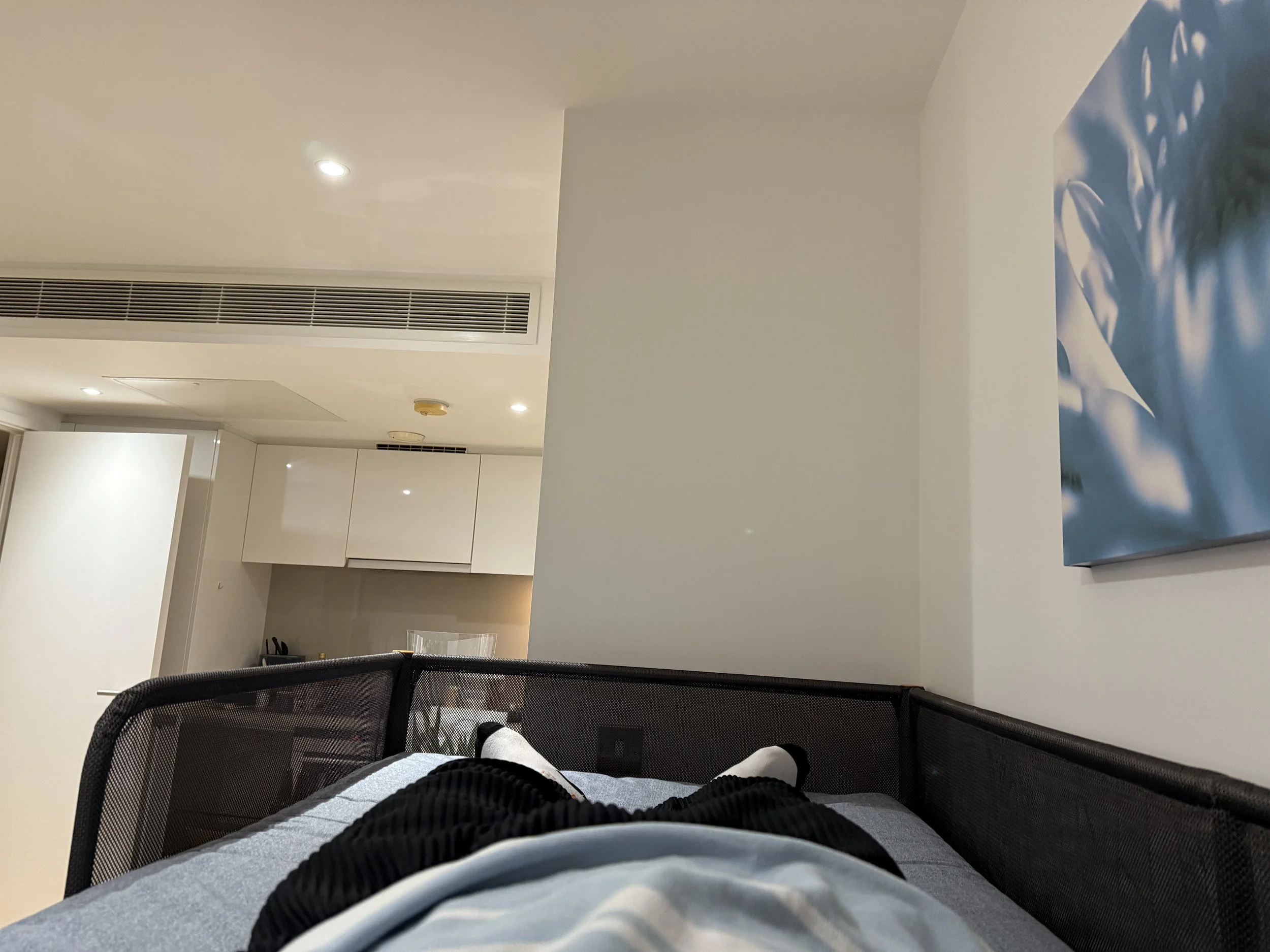 First-person view of a person lying on a bed with gray and black striped pajamas, in a room with white walls, ceiling lights, a blue and white wall art, and a black mesh backing of a chair or crib in the foreground.