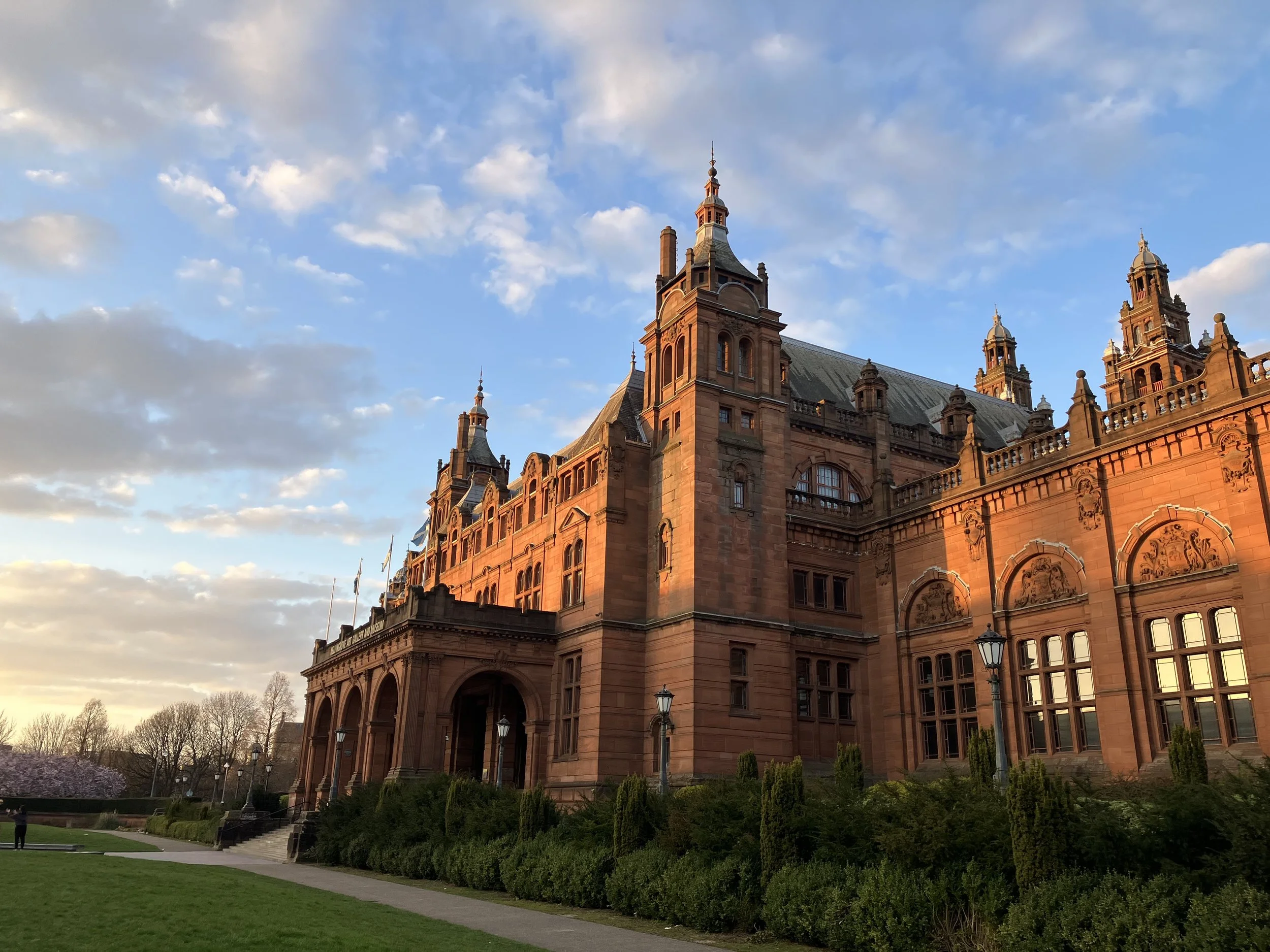 A historic red sandstone castle with multiple towers and ornate architecture, surrounded by greenery and a pathway.