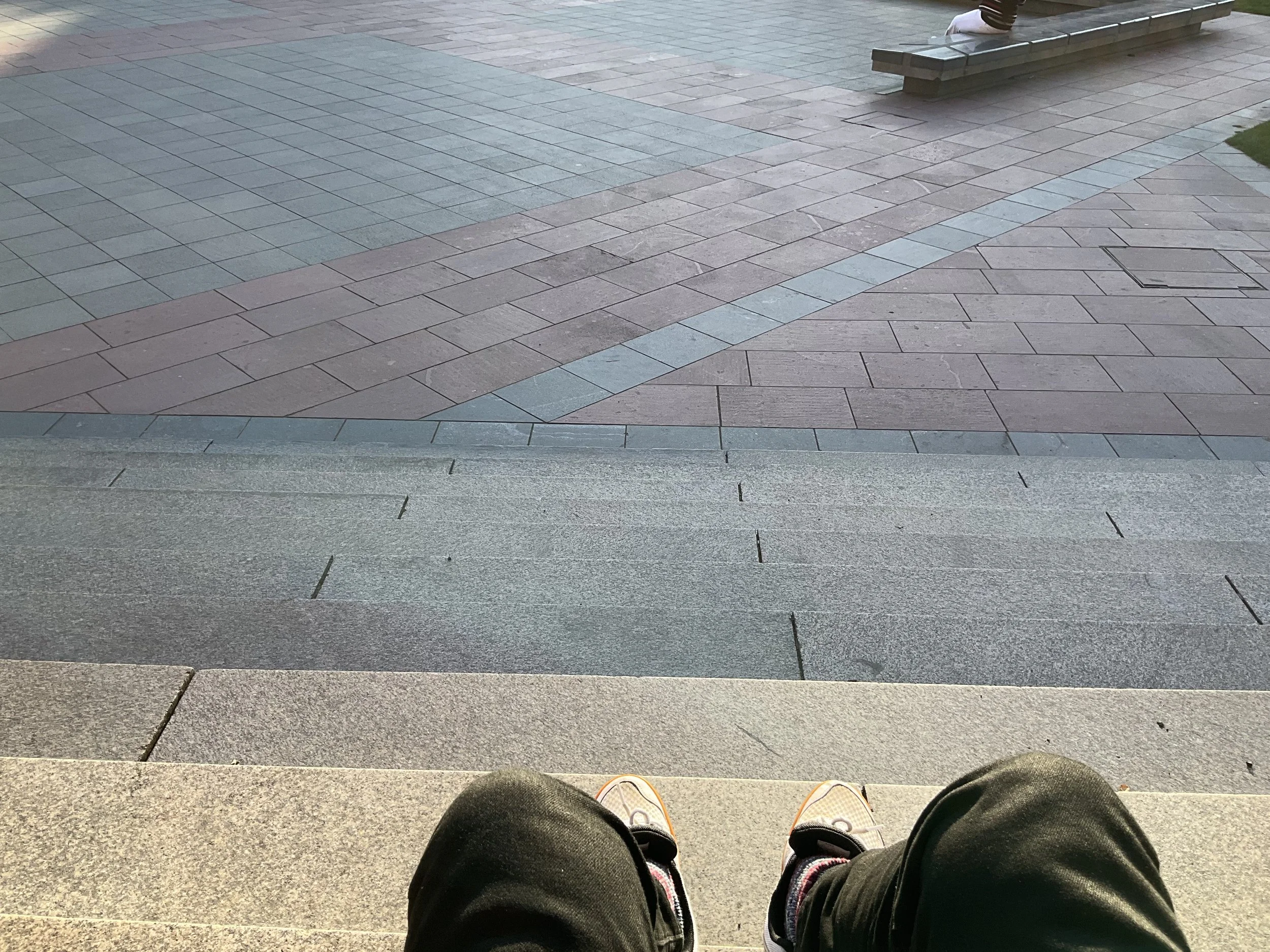 View of a paved area from a person's perspective sitting with feet visible, showing different colored tile patterns on the ground and some grass in the corner.