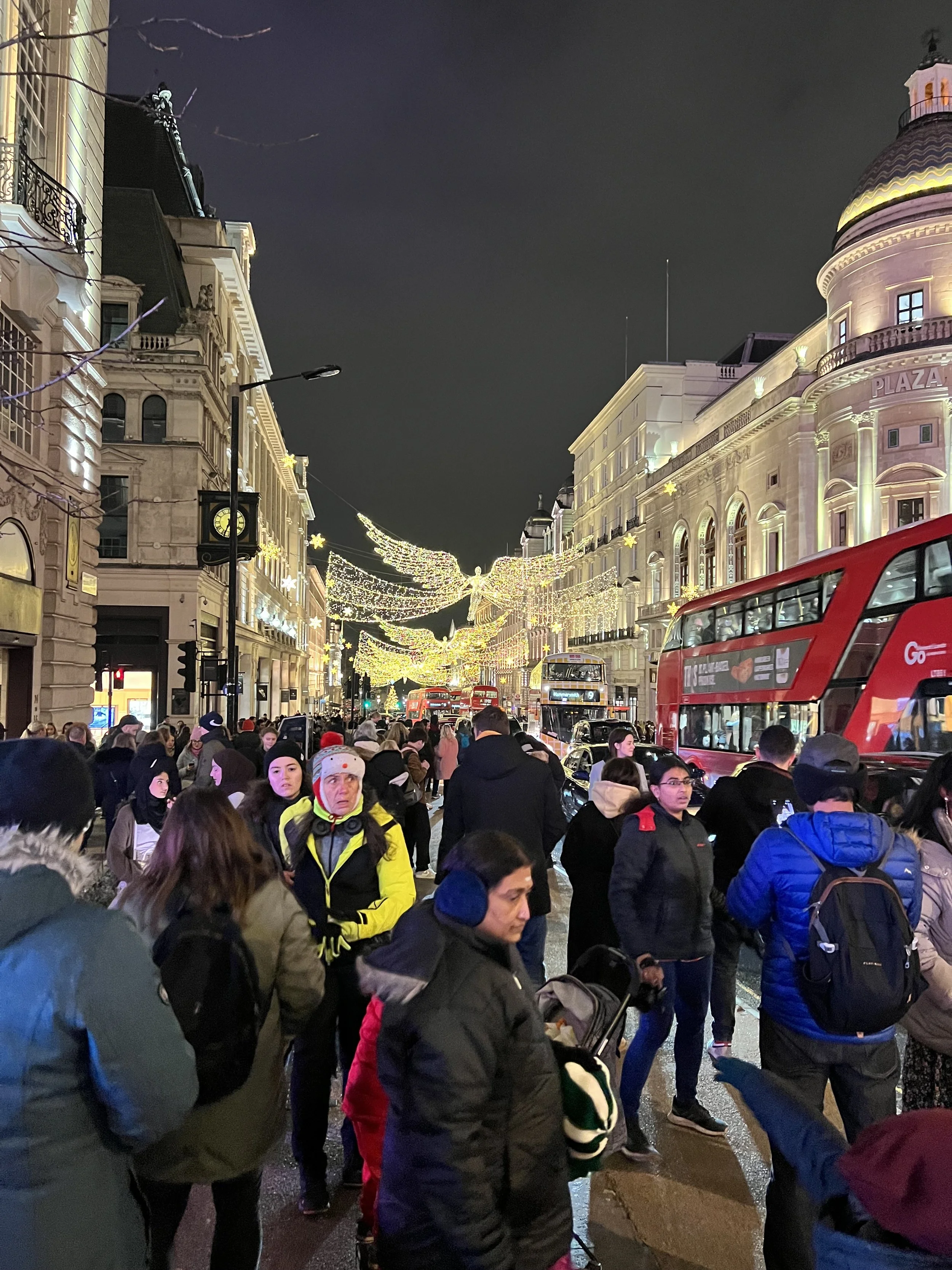 Crowd of people walking on a city street decorated with holiday lights and angel decoration overhead at night, double-decker buses passing by.