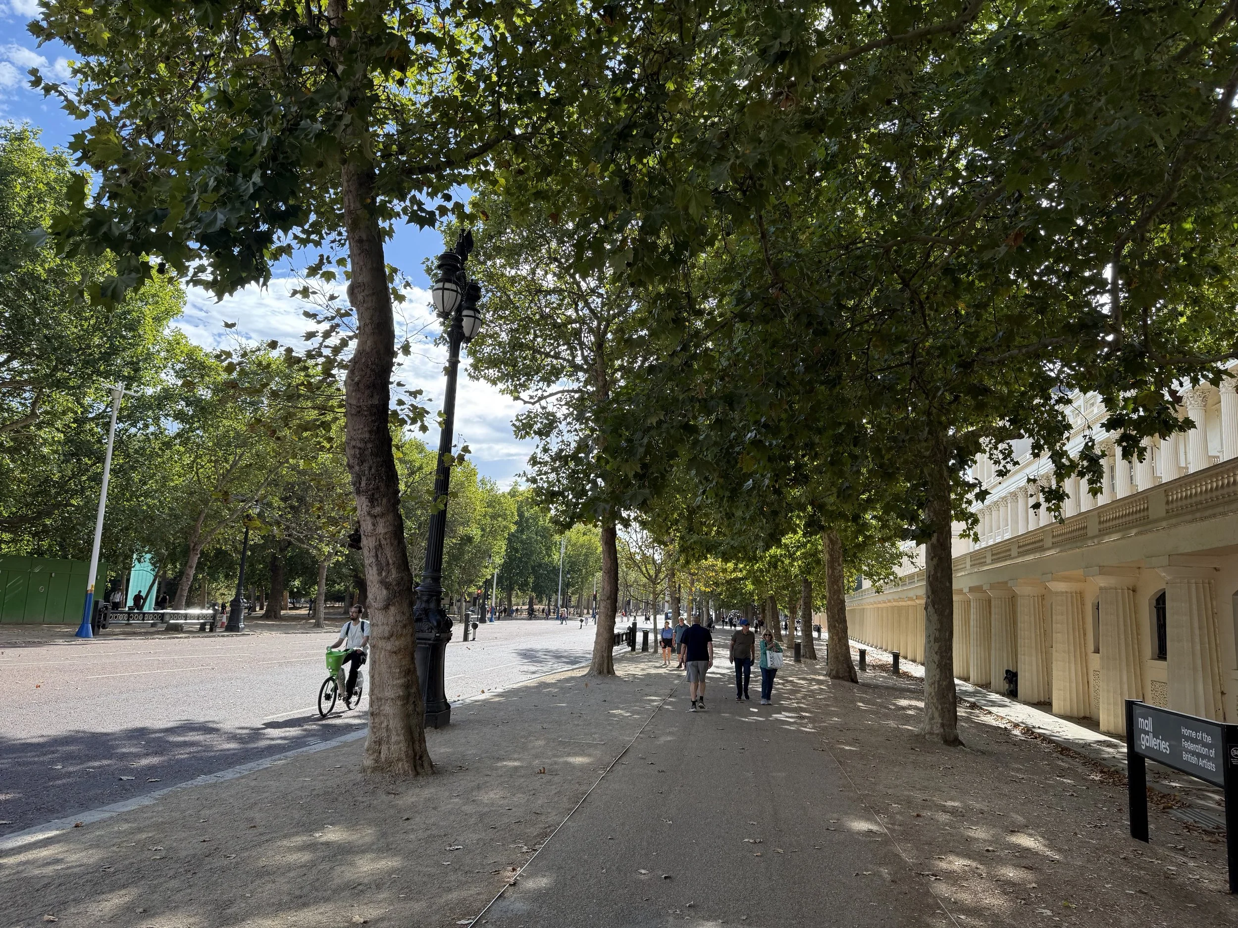 A park with tall trees, a walking path, and people walking and biking. There are lampposts and a building with columns on the right side. The sky is partly cloudy.