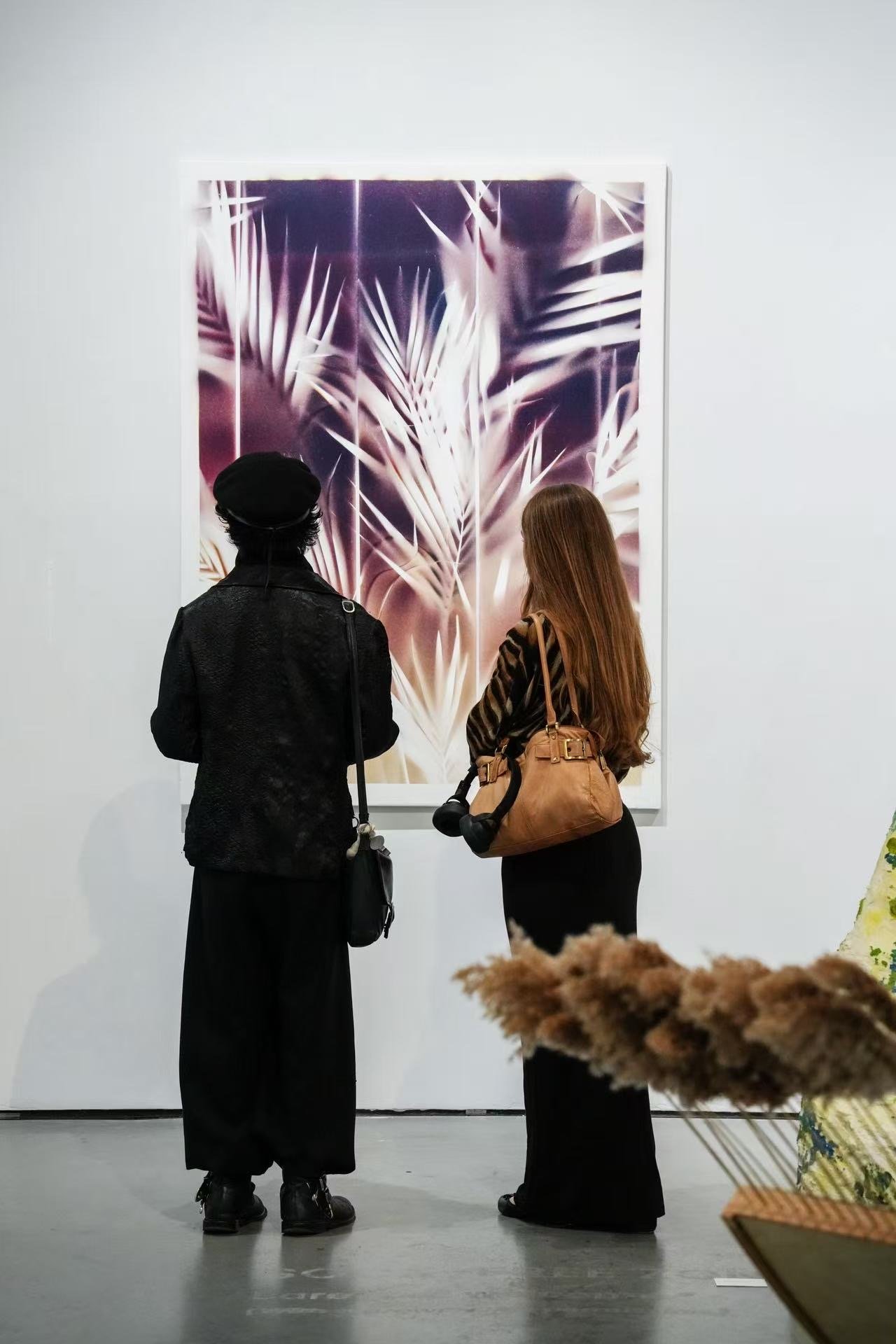 Two women with long hair, one wearing black and the other with brown hair, looking at a large artwork with abstract palm leaves in a gallery.