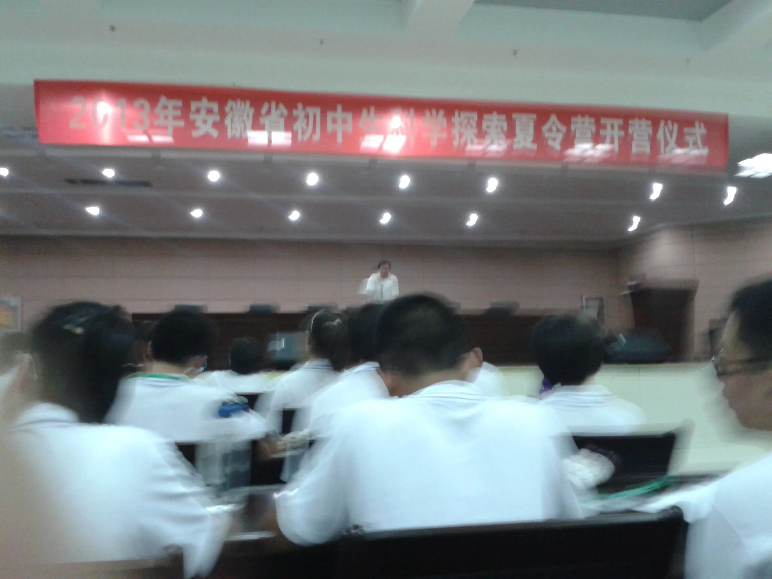 A large group of students, mostly dressed in white shirts, are seated in an auditorium or lecture hall attending an event or ceremony. There is a stage with a woman standing at the podium, and a red banner with Chinese writing hanging above the stage