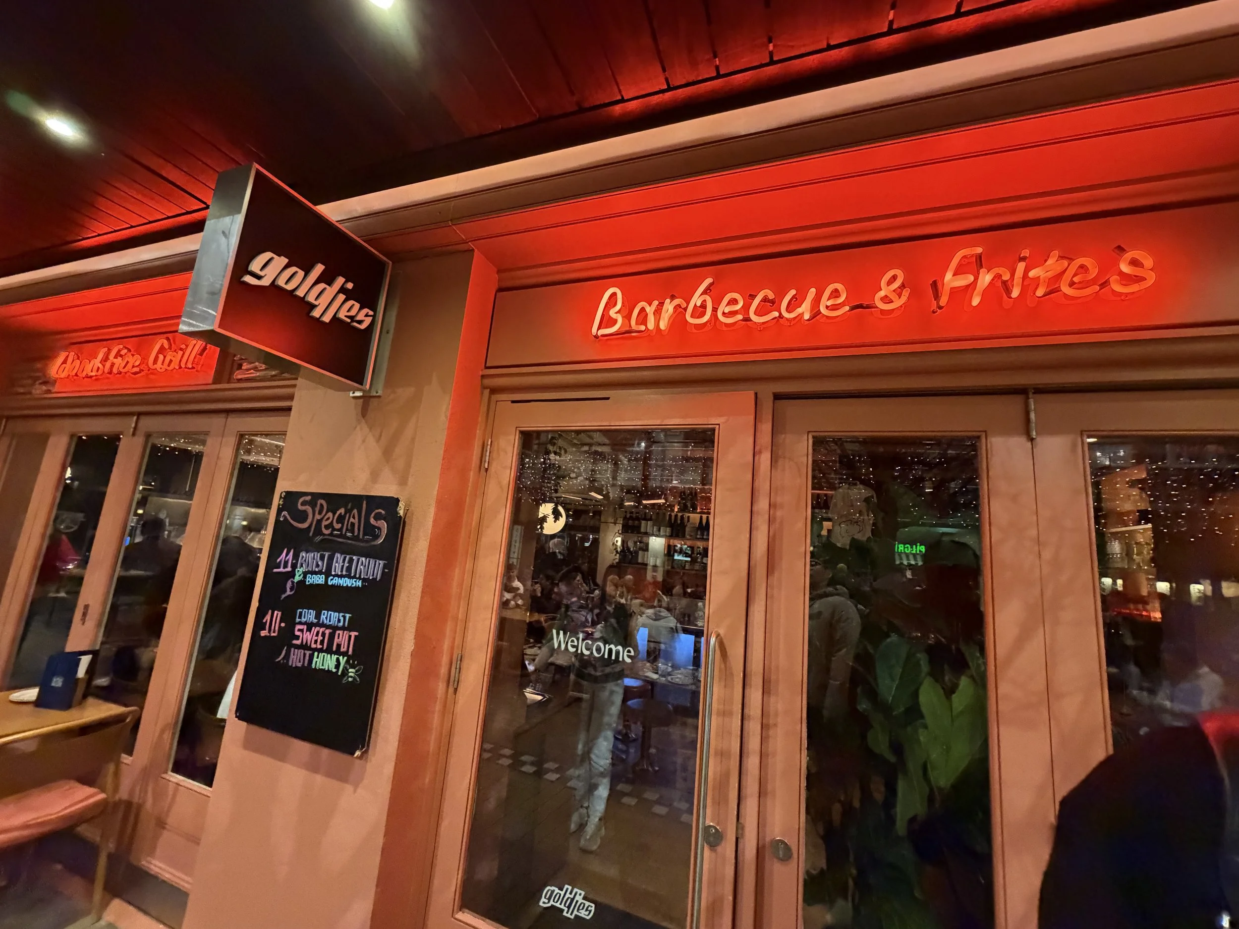 Restaurant facade with a neon sign that says "Barbecue & Fries" and a blackboard menu outside listing various food items, with glass doors and windows revealing diners inside.