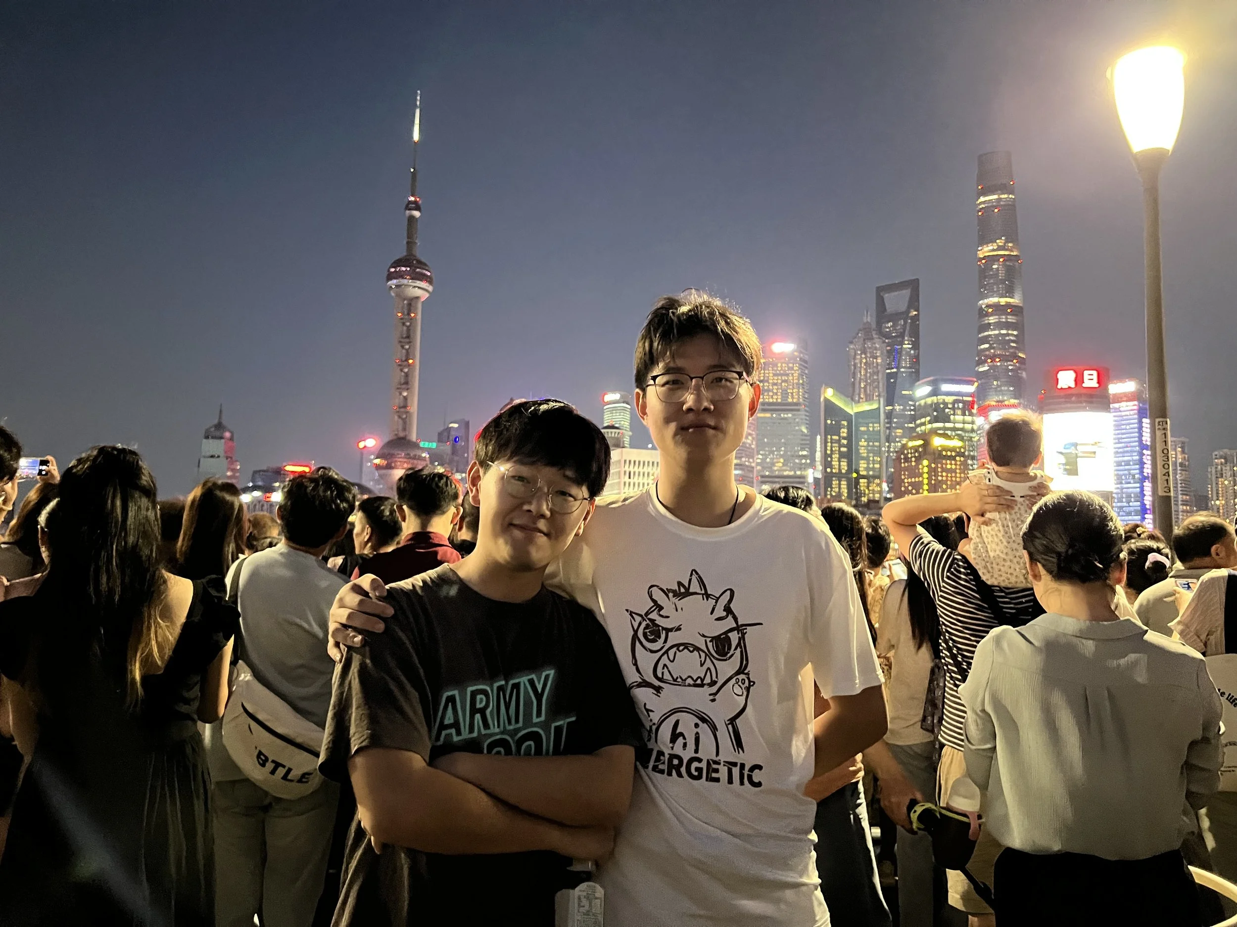 Two young men stand among a crowd of people on a city street during the evening, with tall skyscrapers and city lights in the background.