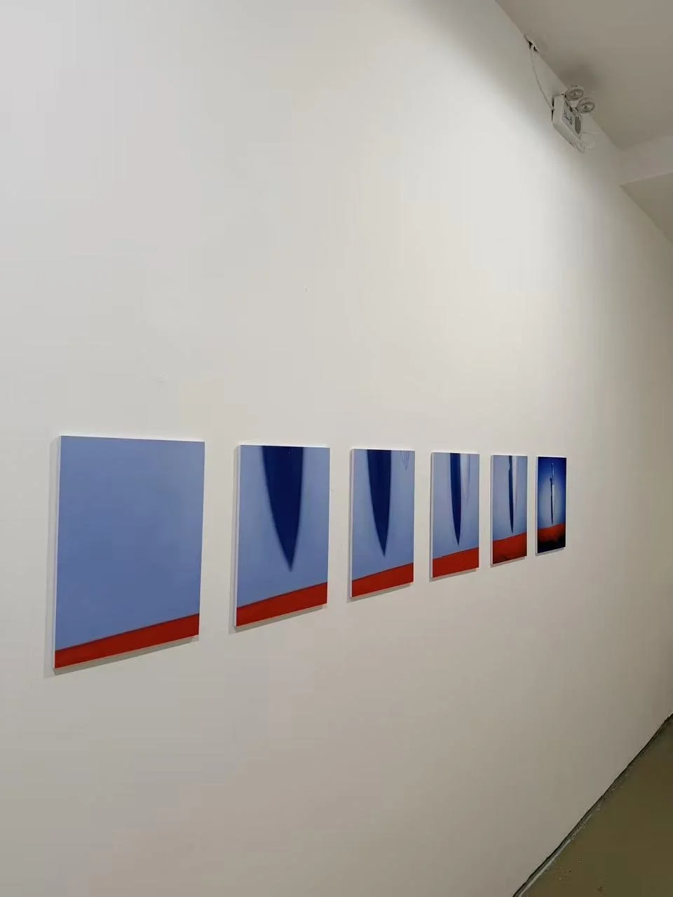 A series of five small abstract paintings with blue backgrounds, red horizontal stripes, and dark cone-shaped forms, hung in a row on a white gallery wall.