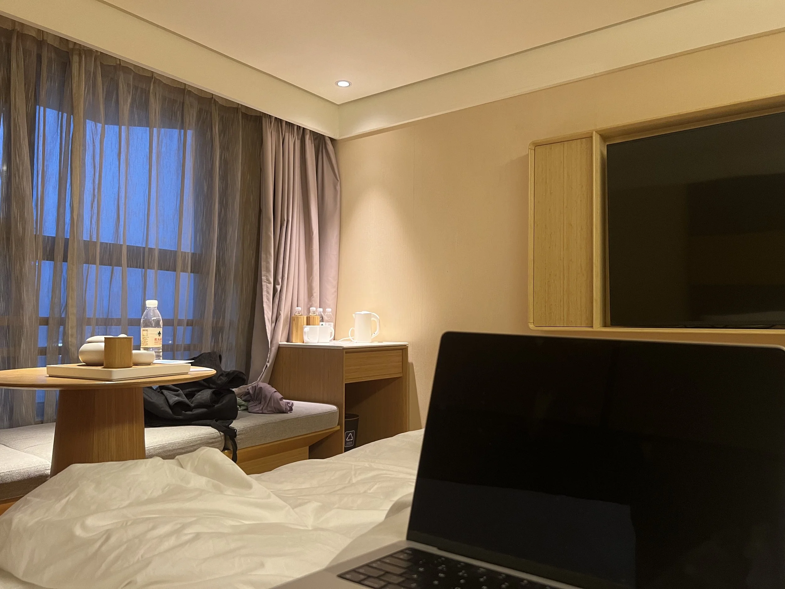 A hotel room with a bed, a table with a water bottle and cups, sheer curtains, and a wall-mounted TV.