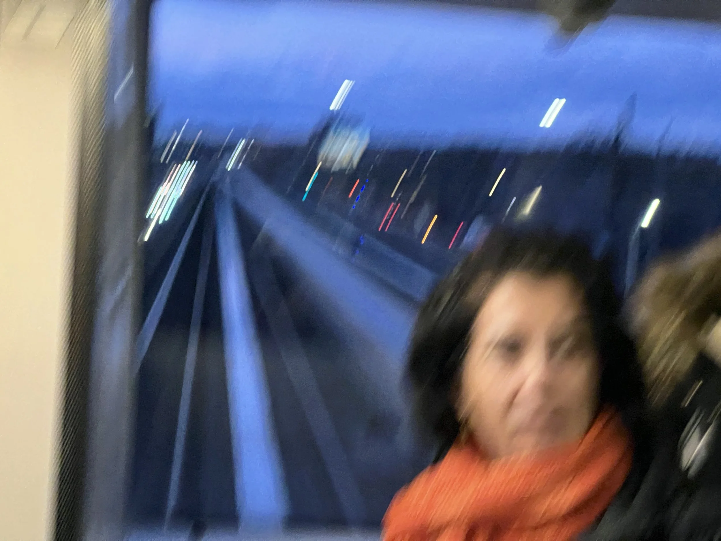 Blurred photo of a young girl with long hair wearing an orange top, standing in front of a blue window or wall, possibly inside a vehicle such as a bus or train.