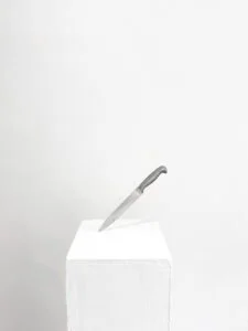 A knife stabbing into a white rectangular pedestal against a plain white background.