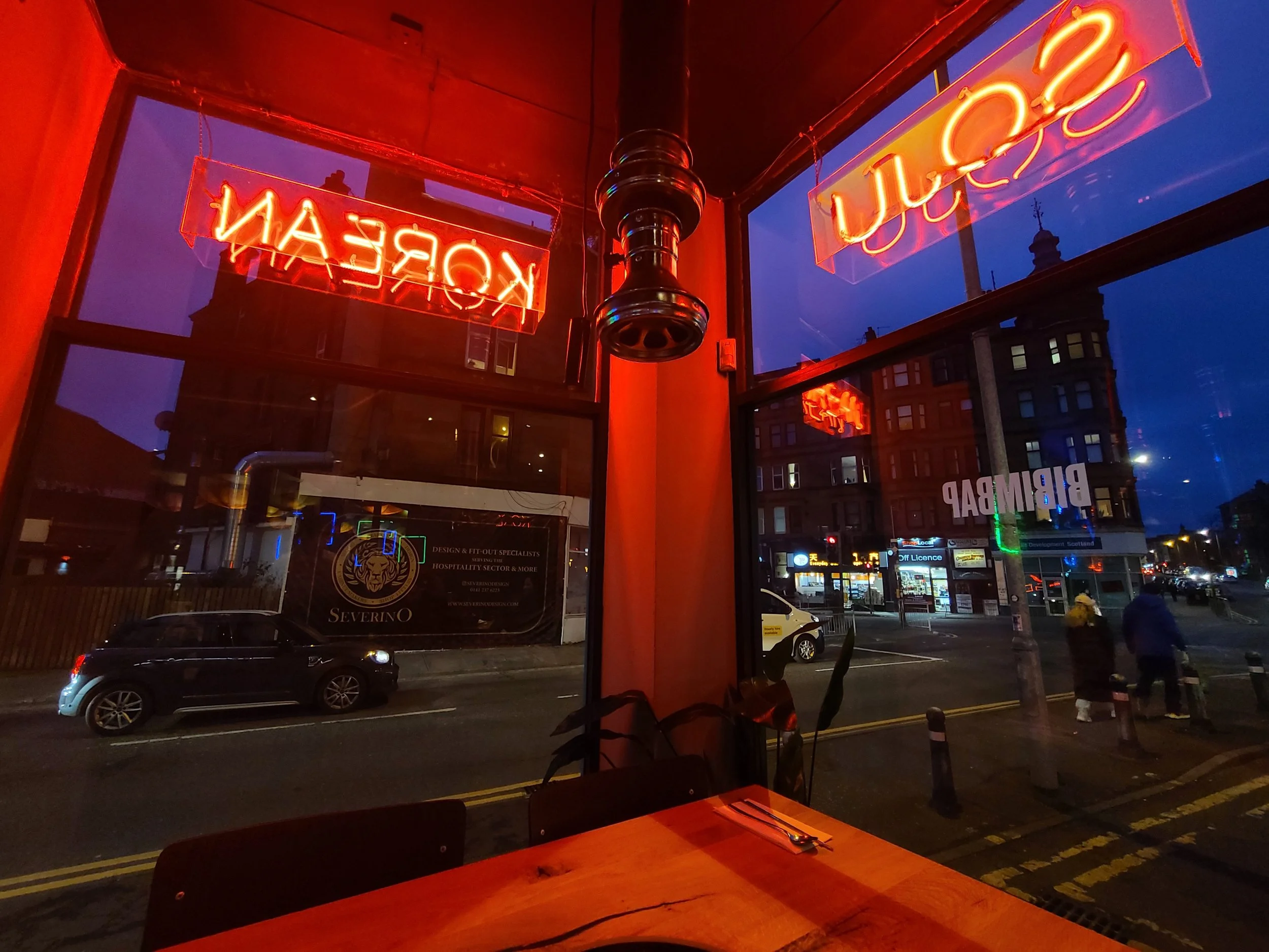 View from inside a cafe or restaurant showing two neon signs outside through the window that read 'OPEN' and 'KOREAN'. There is a street with cars and pedestrians, and buildings with lit windows in the background. The scene is during the evening or n