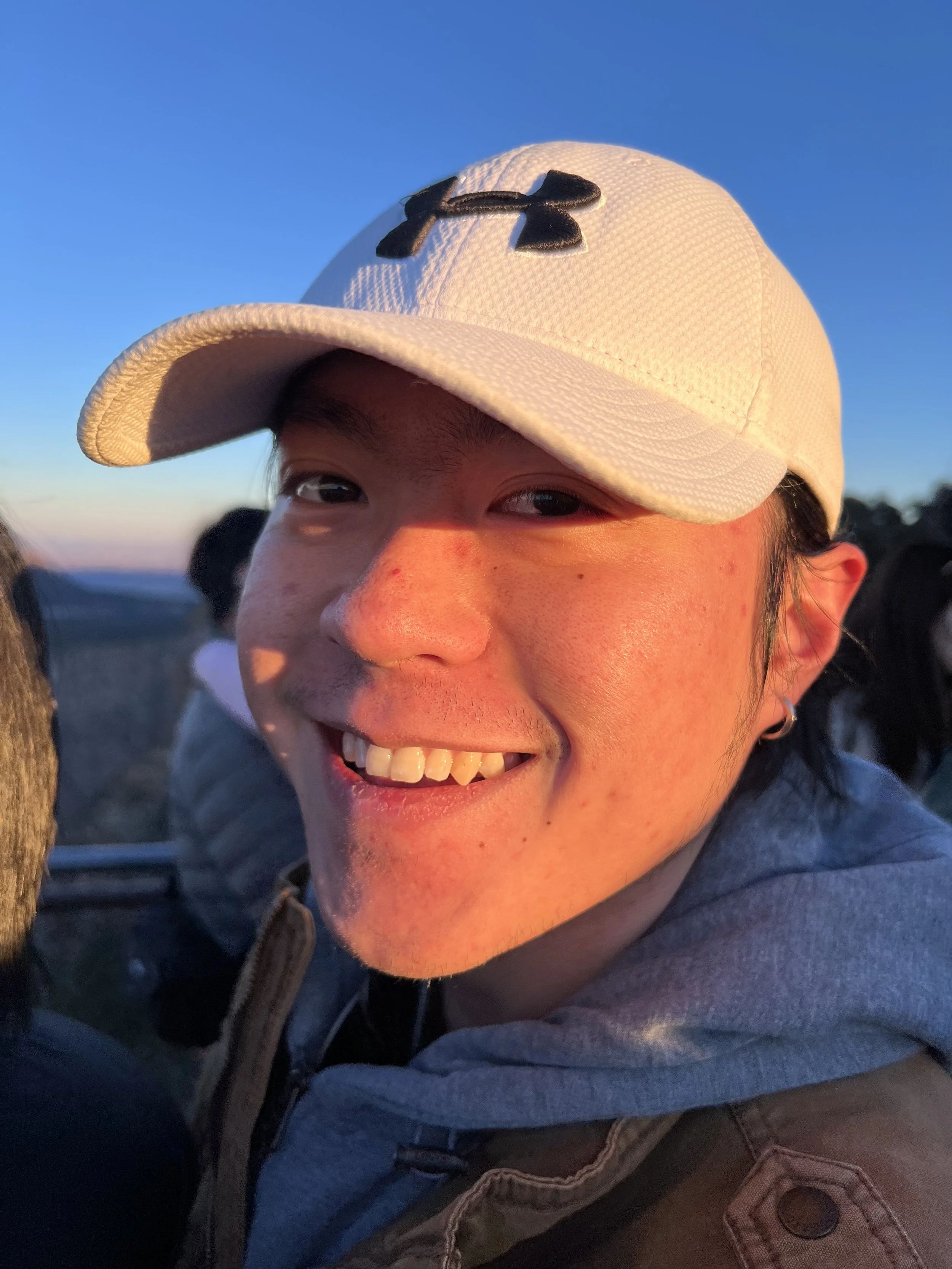 The image shows a close-up of a smiling person wearing a white Under Armour baseball cap, with a clear blue sky in the background.