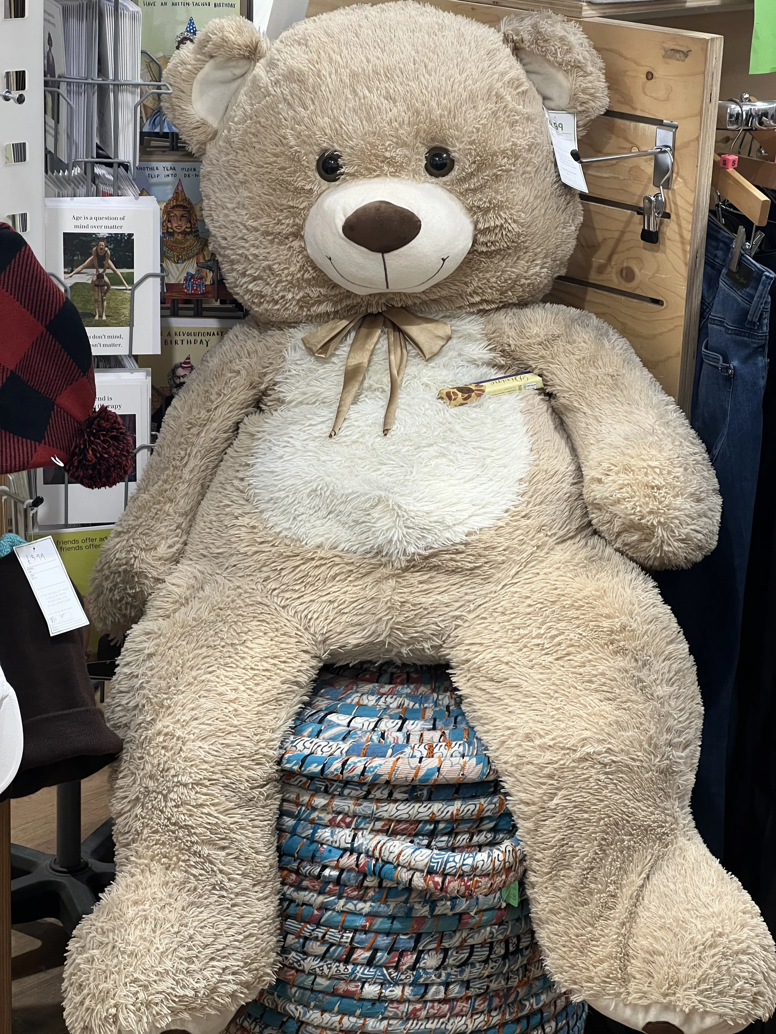 A large, plush teddy bear with tan fur, a white muzzle, and a brown nose, sitting on a display shelf in a store surrounded by other items including stuffed animals, books, and clothing.
