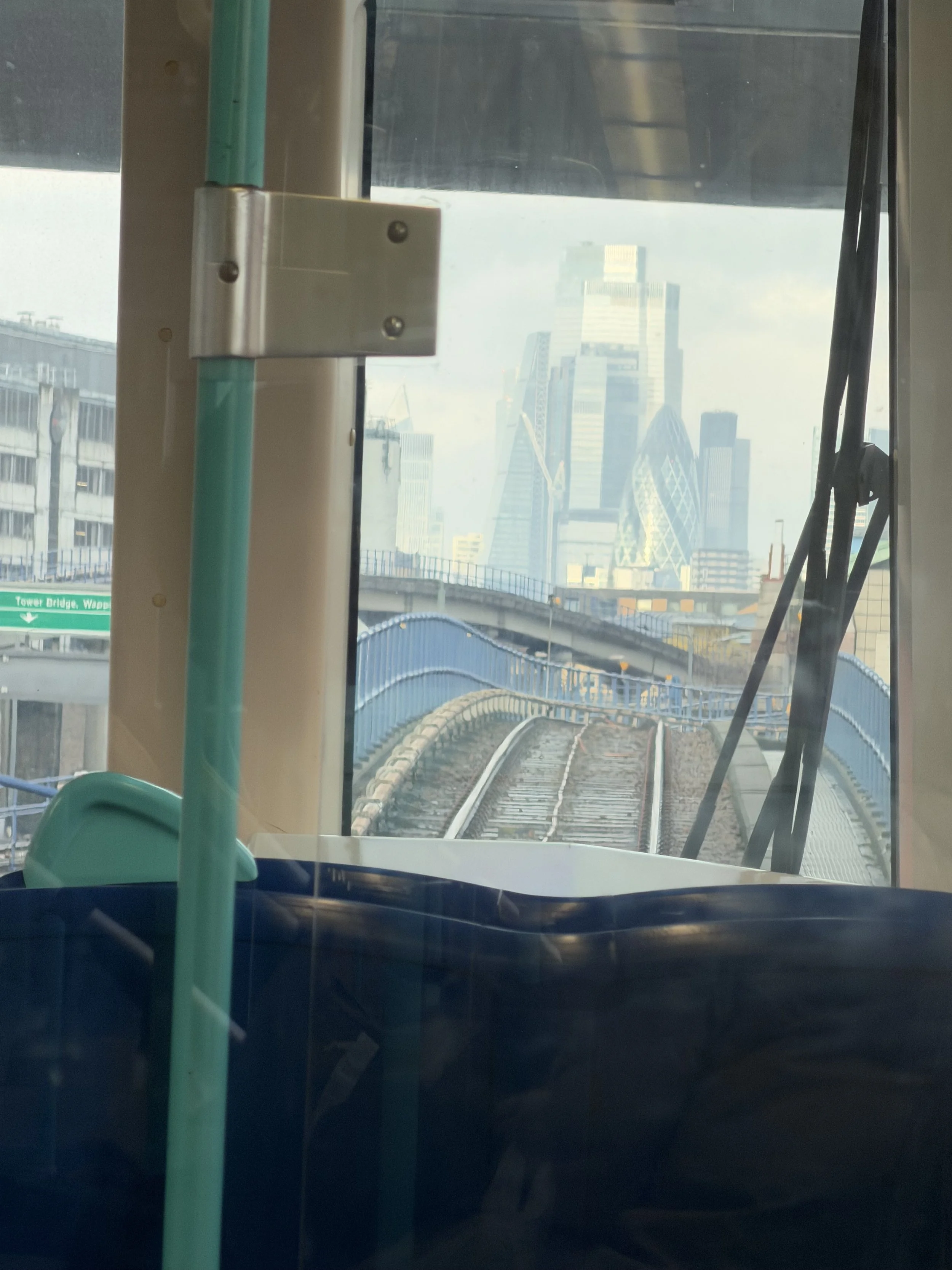 View from inside a train looking out through a window at a city skyline with tall modern buildings, a bridge, and a sign that reads 'Tower Bridge, Wapping'.