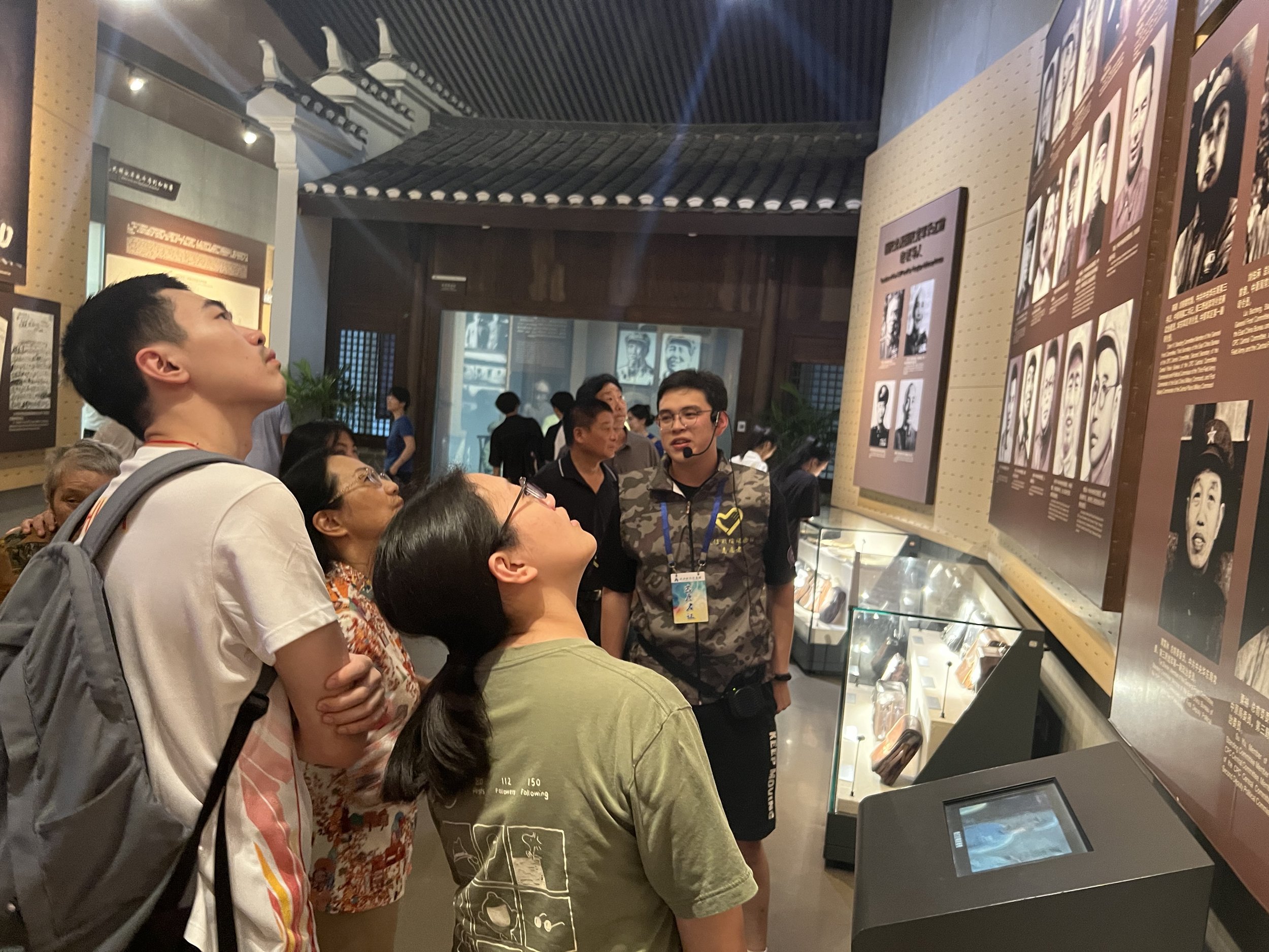 People at a museum exhibit looking at displays with photographs and text. A young man with a backpack is in the foreground, looking up. Other visitors are gathered around the exhibits.