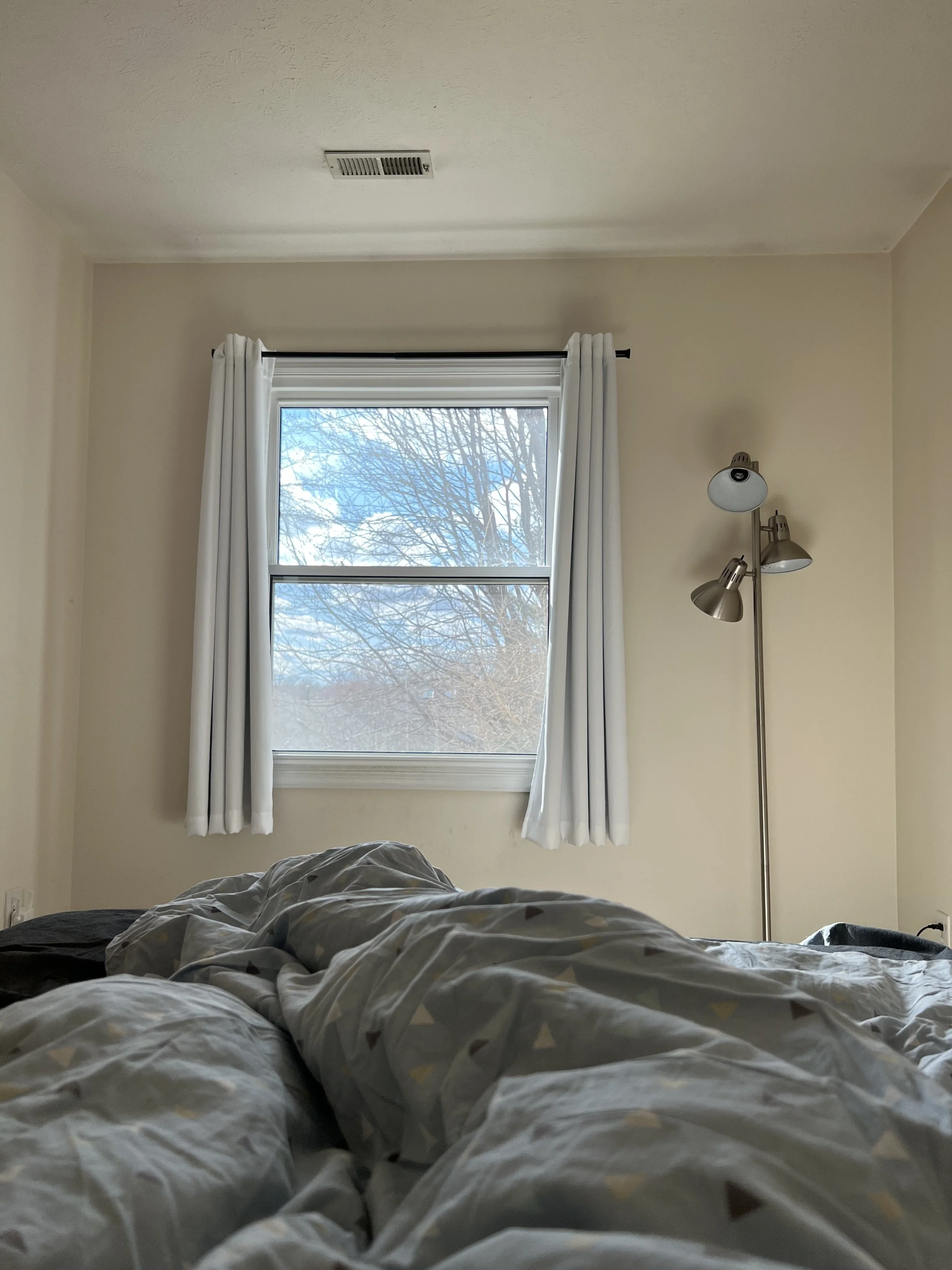 A bedroom with a large window covered with white curtains, a ceiling air vent, a floor lamp with multiple adjustable heads, and a messy bed with patterned bedding.