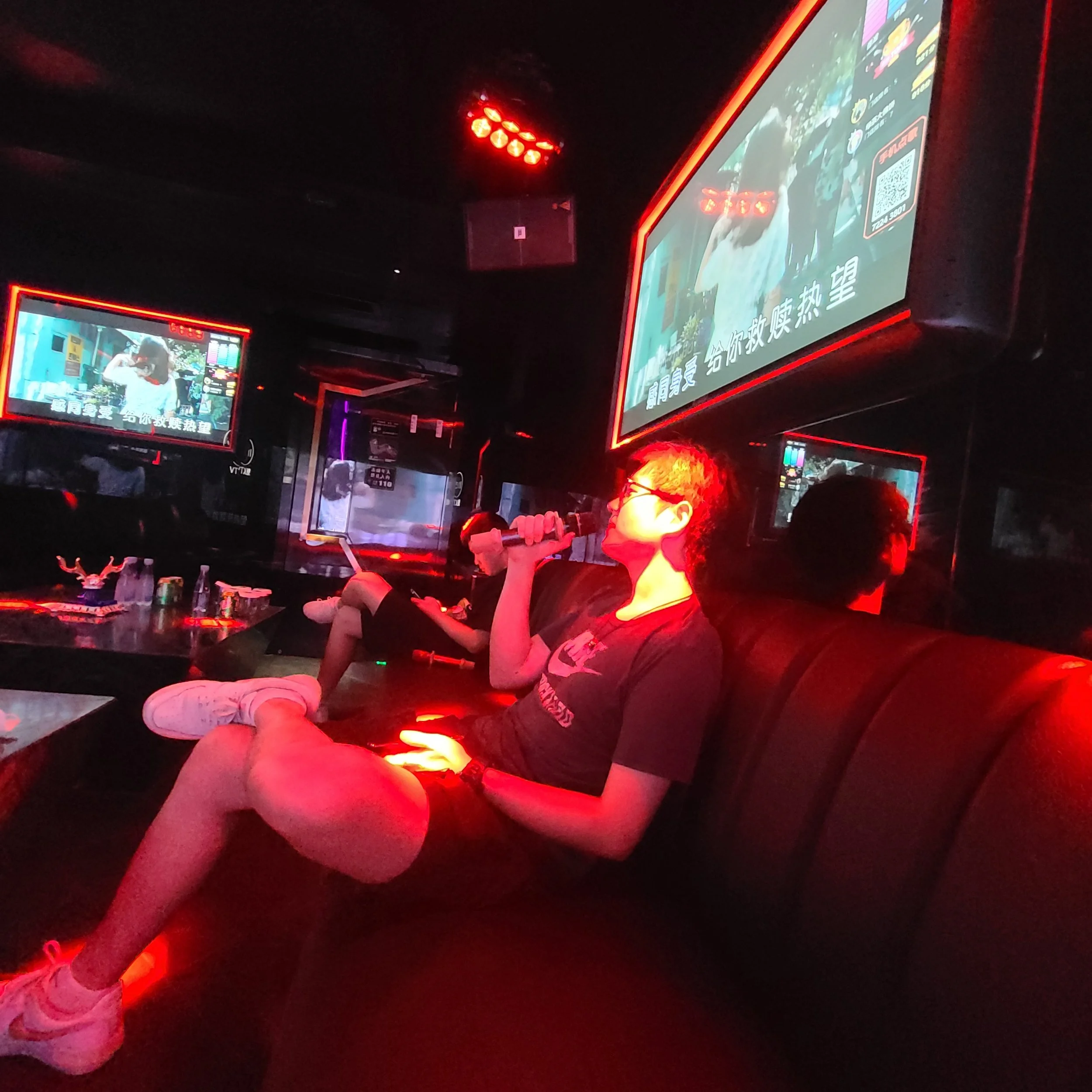 Dark room illuminated by neon red lights, with several LED screens displaying colorful images and text in Chinese. A person stretching or exercising, wearing white sneakers and beige pants, is visible in the foreground.