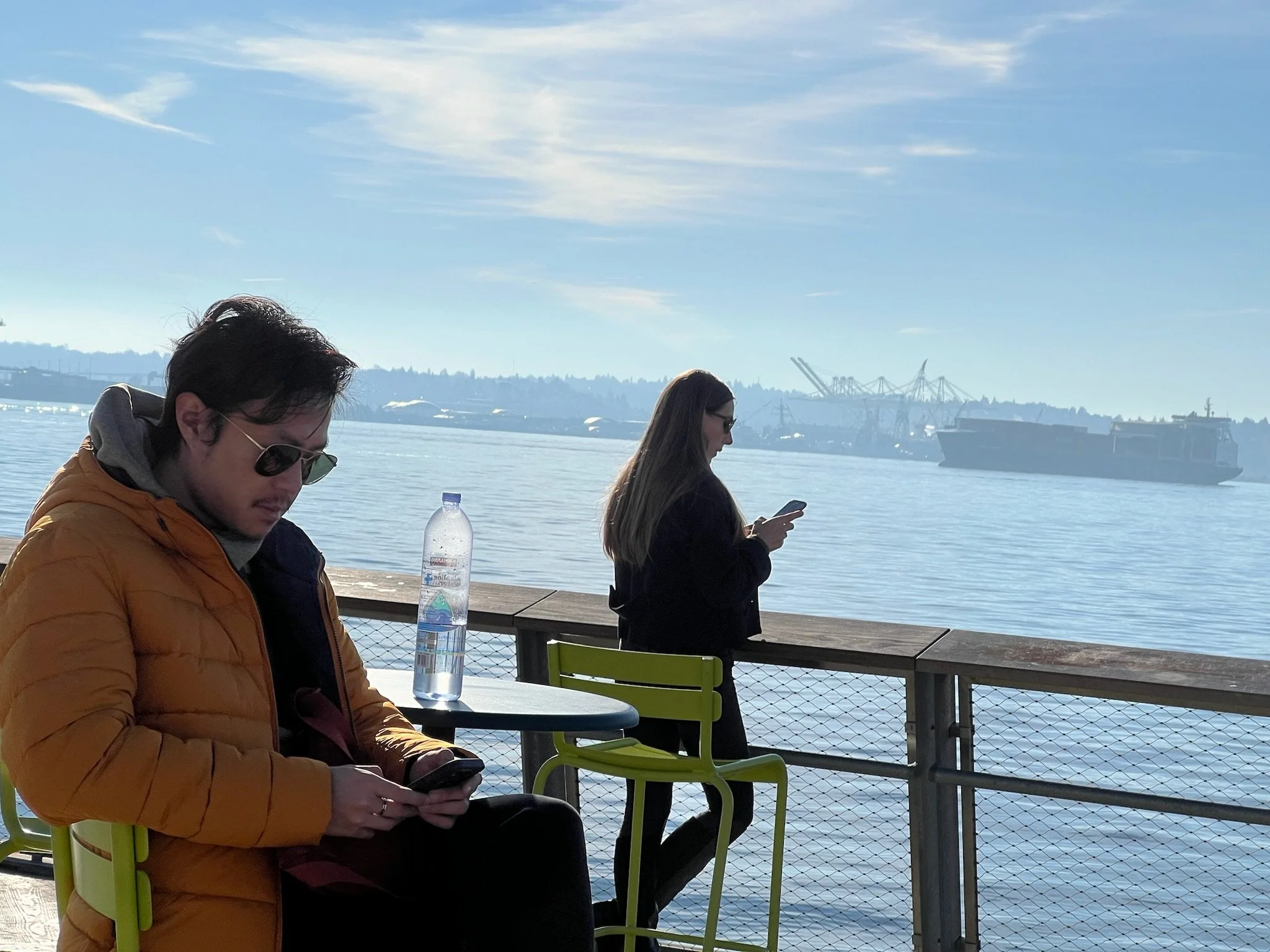 Two people sitting at a table near a waterfront, one looking at a phone and the other also looking at a phone, with ships and industrial cranes in the background under a blue sky.