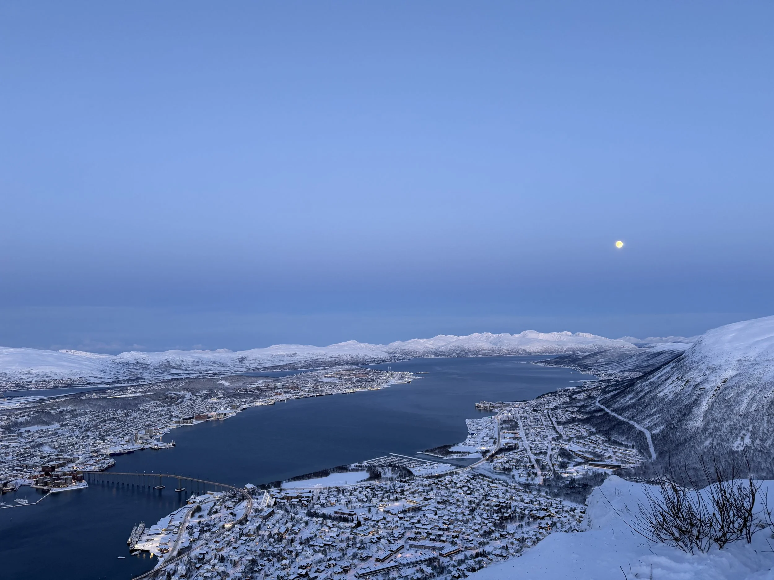 Aerial view of a snow-covered city near a body of water, surrounded by mountains under a clear blue sky with a visible moon.