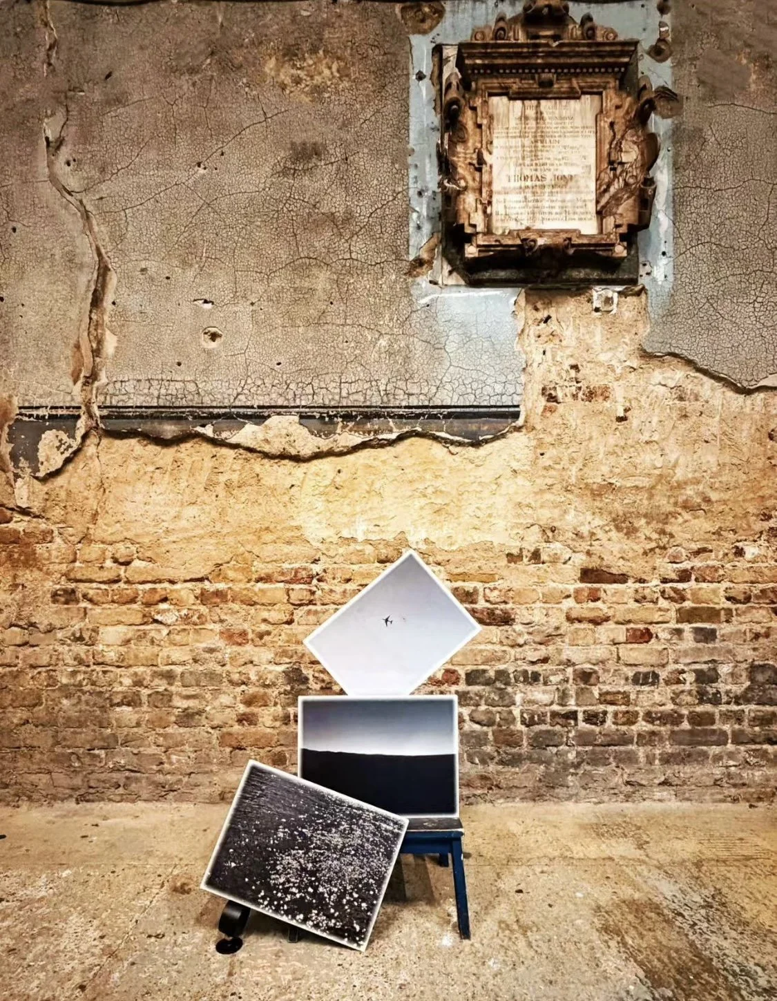 An art installation features three framed photographs and a drone in a rustic space with a textured, cracked wall and exposed brick. The photographs are on a small chair, with the drone flying above them.