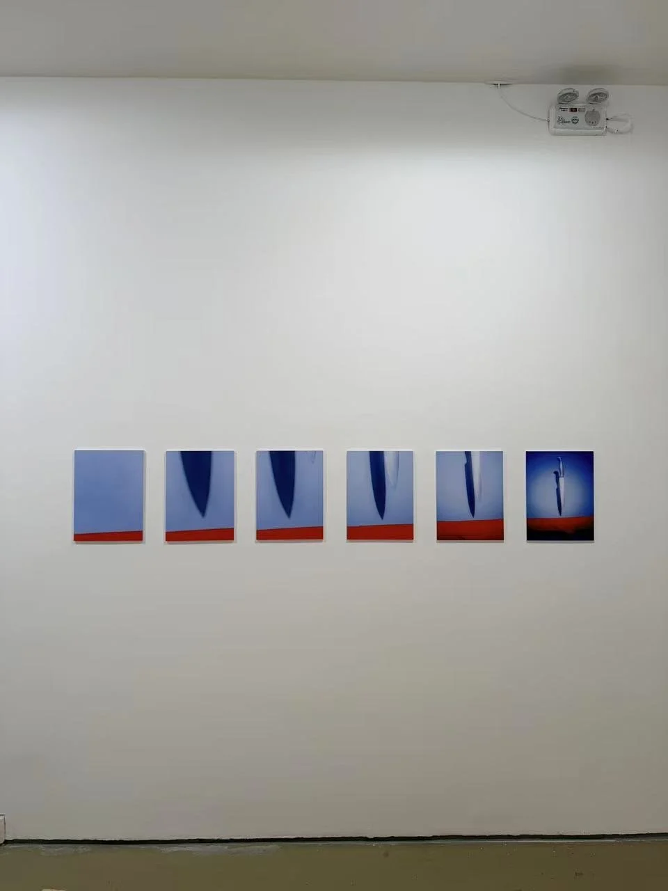 Six small hotel room paintings in a row on a white wall, each depicting a blue sky with a black umbrella opened and closed, with a red strip at the bottom. There is an electrical panel in the upper right corner.