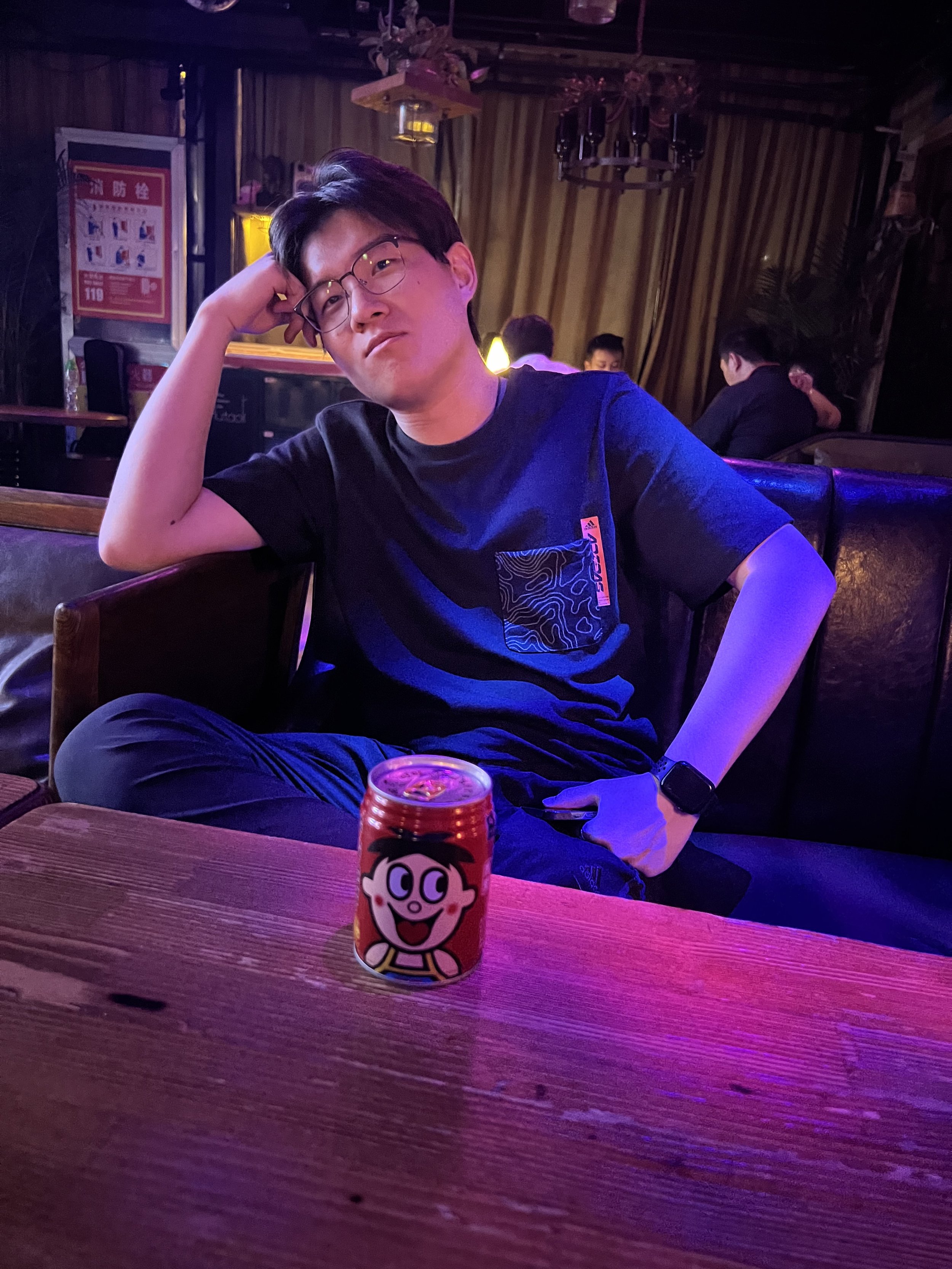 A young man with glasses is sitting in a dimly lit room, resting his head on his hand, with a relaxed expression. He is wearing a dark t-shirt and has a smartwatch on his left wrist. In front of him on a wooden table, there is a small can of characte