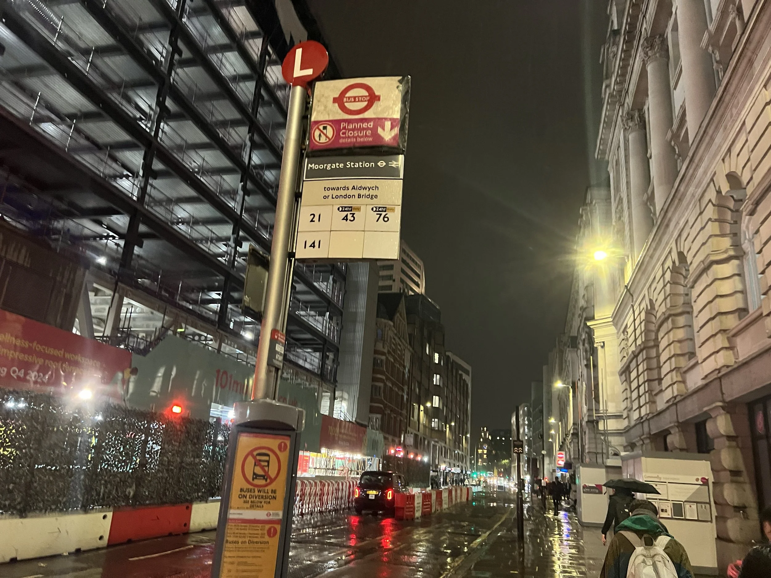 Nighttime city street scene with a bus stop sign indicating planned closure, traffic, and streetlights reflecting on wet pavement, with nearby buildings and sidewalk.