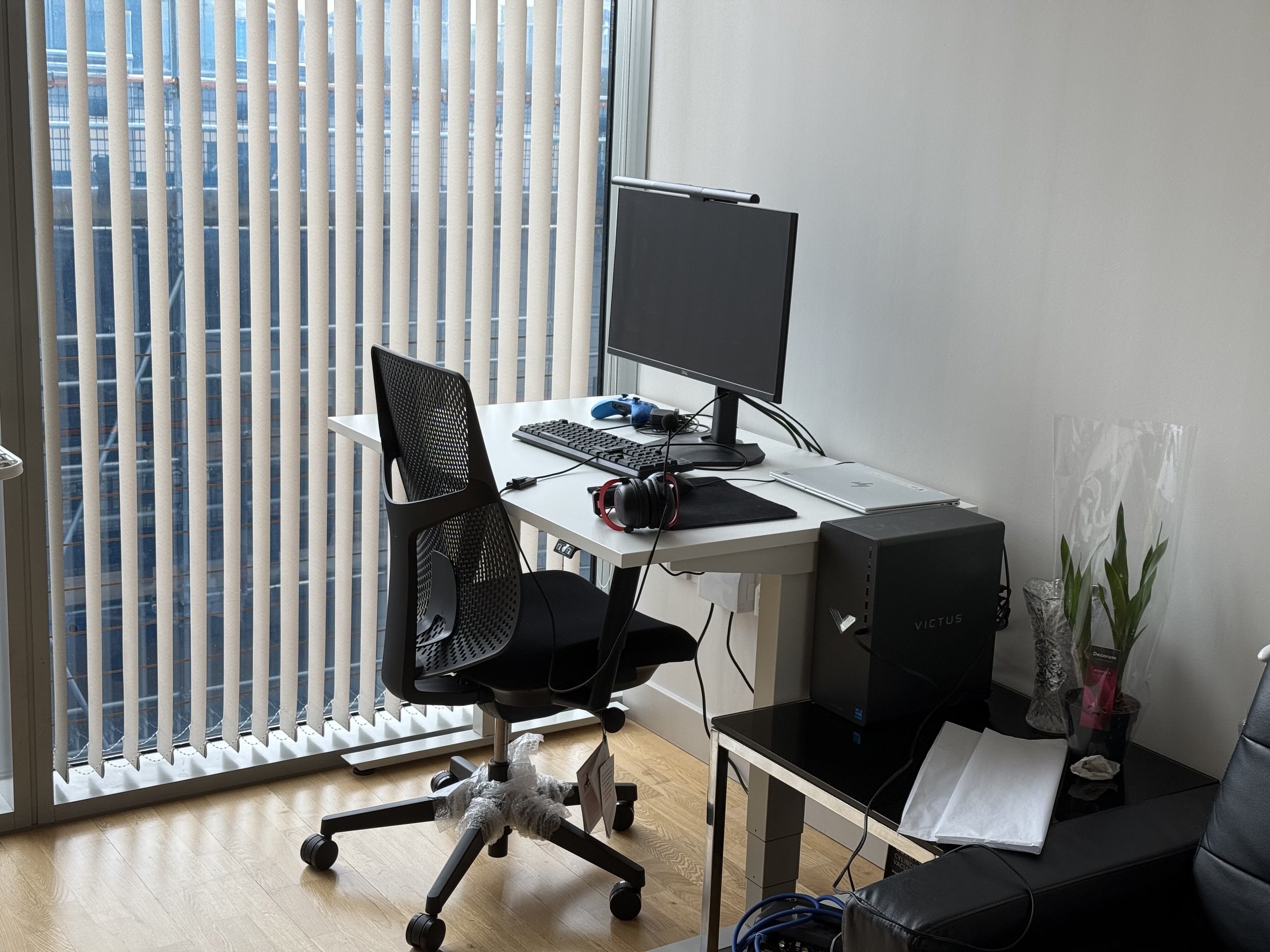 A small office space with a white desk, black office chair, computer monitor, keyboard, and gaming headset. There is a window with vertical blinds, and a plant in a pot near the desk. A black computer tower is on a small table next to the desk.