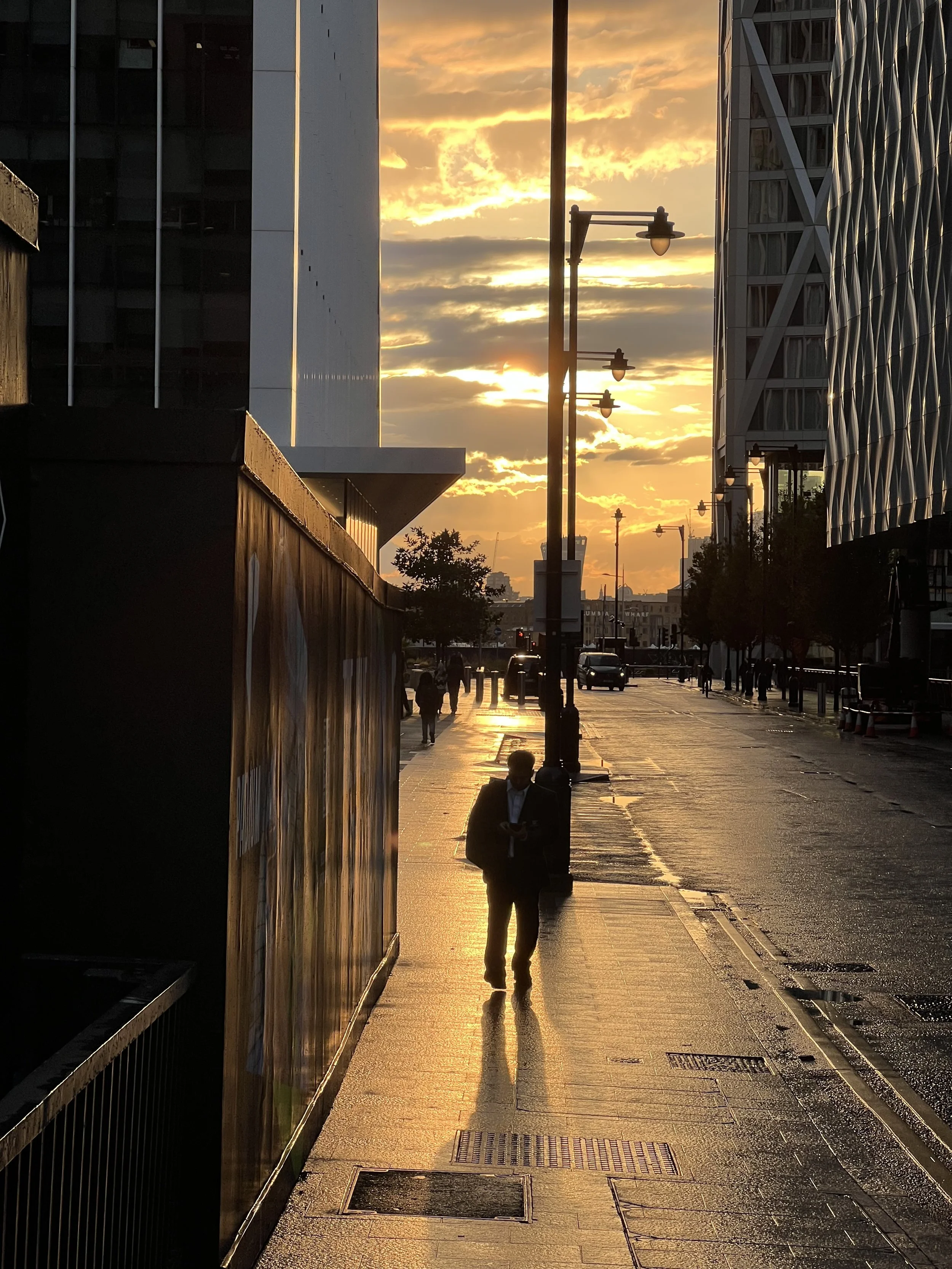 A city street at sunset with the sky painted in orange and yellow hues. Silhouettes of buildings, pedestrians, and street lamps are visible, with the sunlight reflecting on the wet pavement.