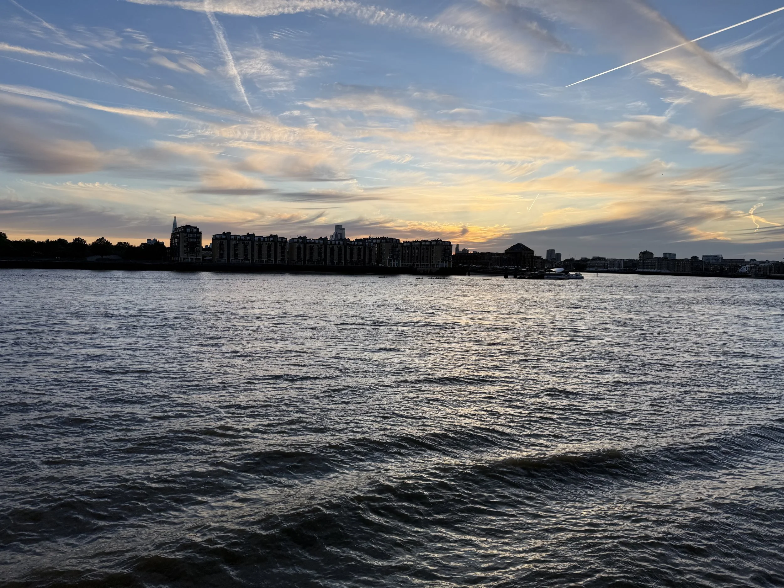 View of a city skyline across a body of water during sunset, with buildings, trees, and a partly cloudy sky with contrails.