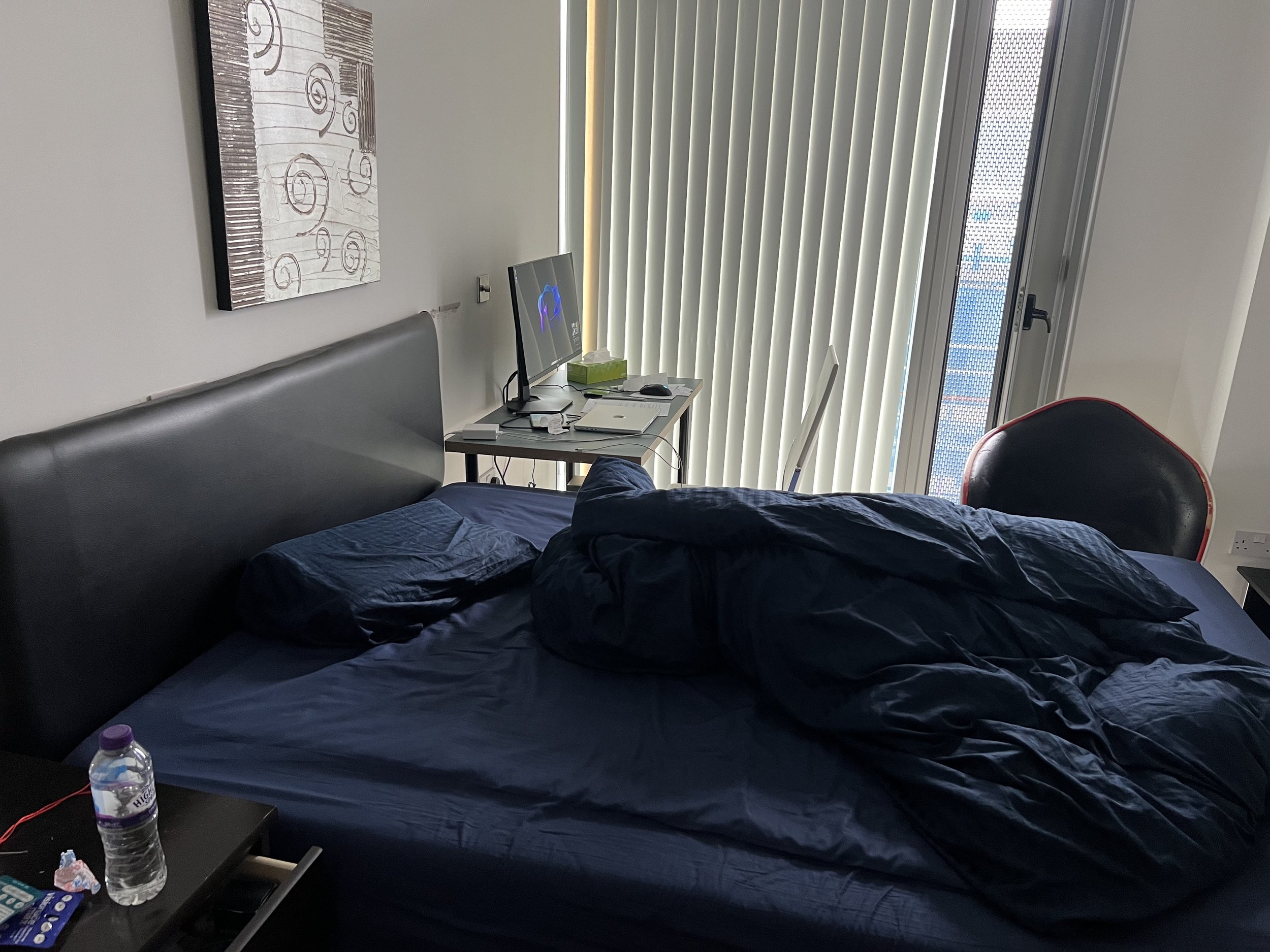 A bedroom with a bed covered in a dark blue blanket, a nightstand with a water bottle, tissues, and remote controls, a mounted computer monitor with glowing lights, a wall hanging with artistic swirls, vertical blinds covering a window or door, and a