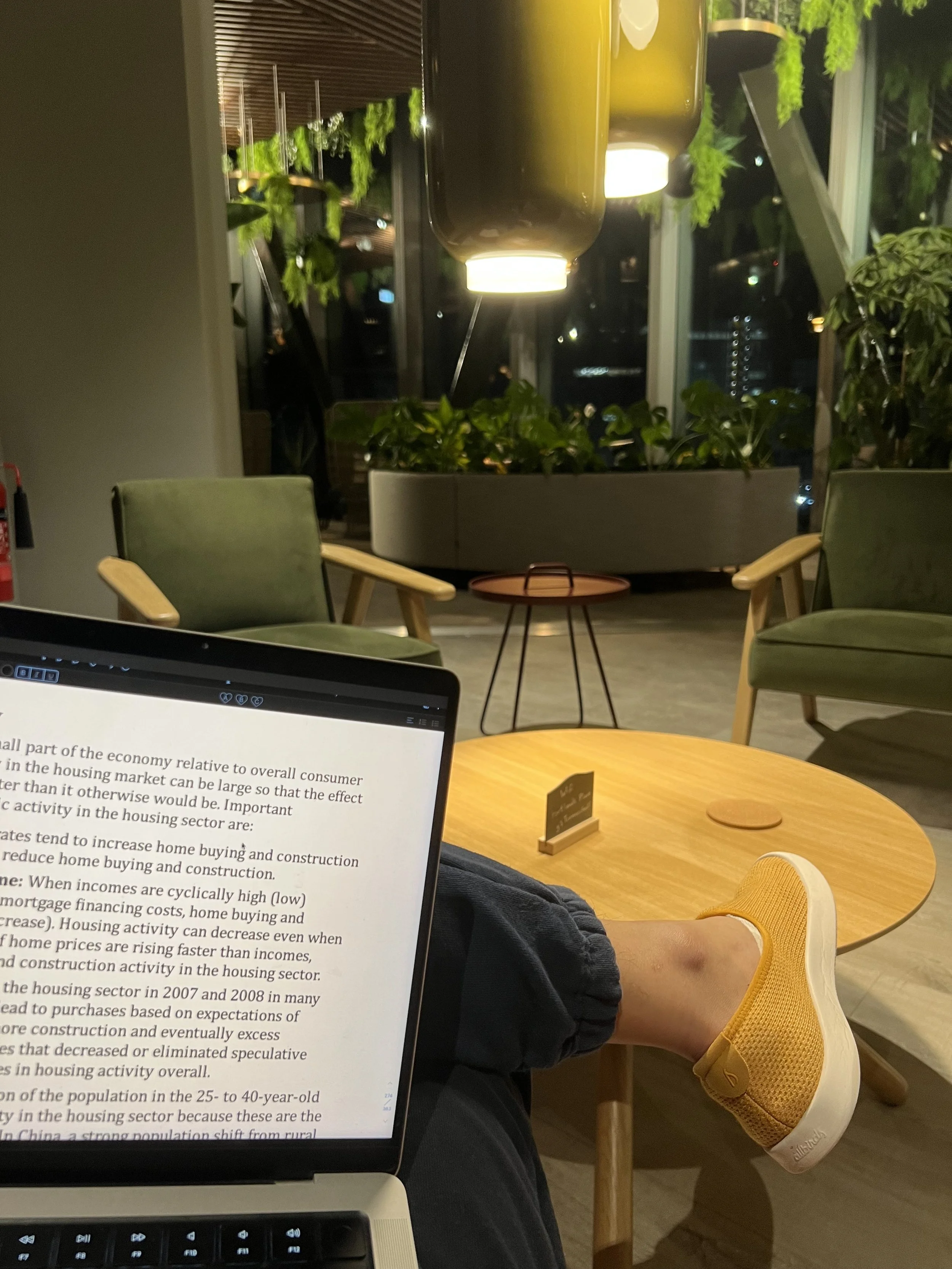 The image shows a person seated at a wooden table in a modern indoor setting, possibly a cafe or office, with a laptop displaying text. The person's yellow sneakers are visible, resting on the floor. In the background, there are large green plants, c