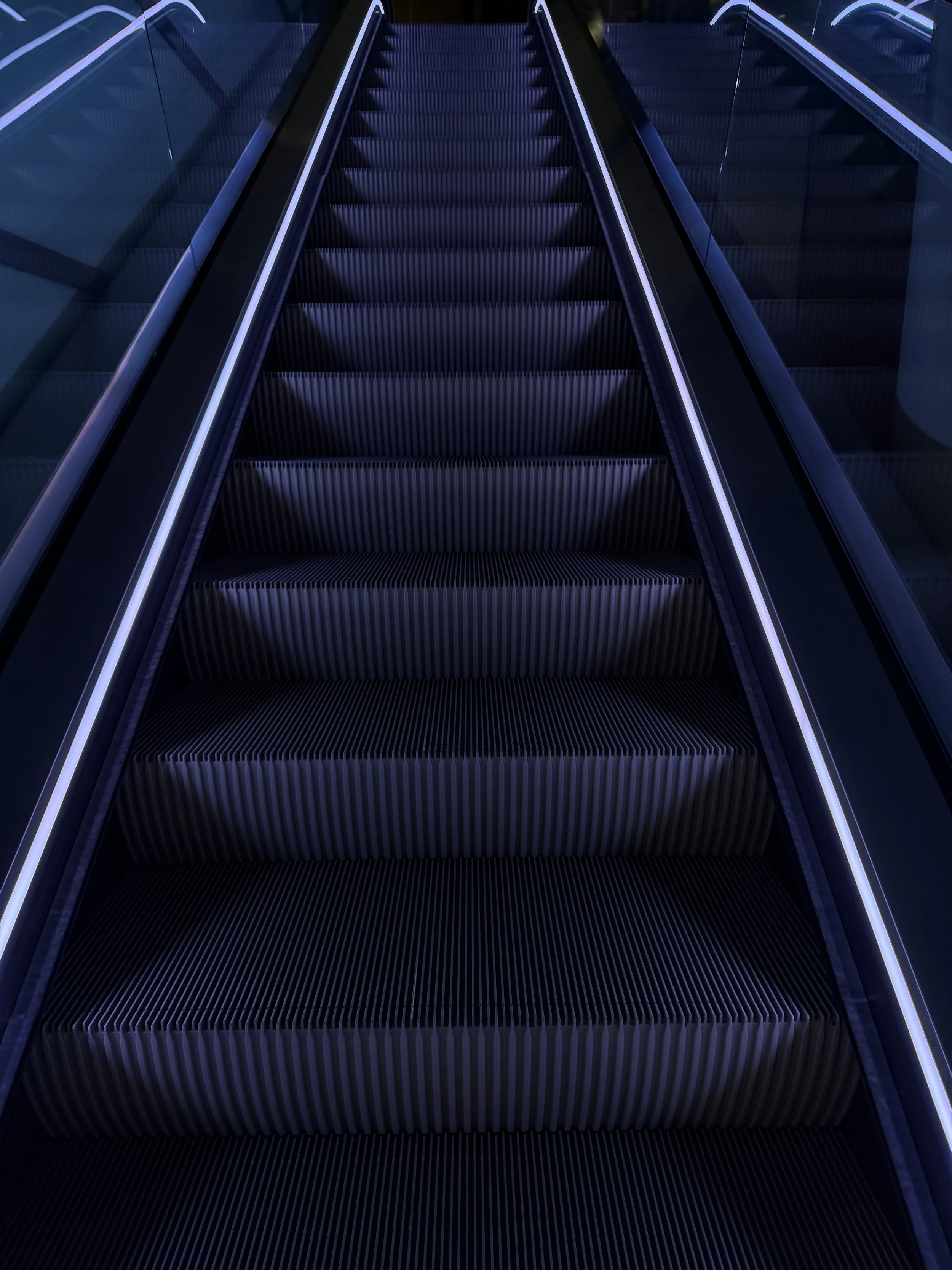 A modern, dark-colored escalator with illuminated handrails and steps, leading upwards in a sleek, futuristic setting.