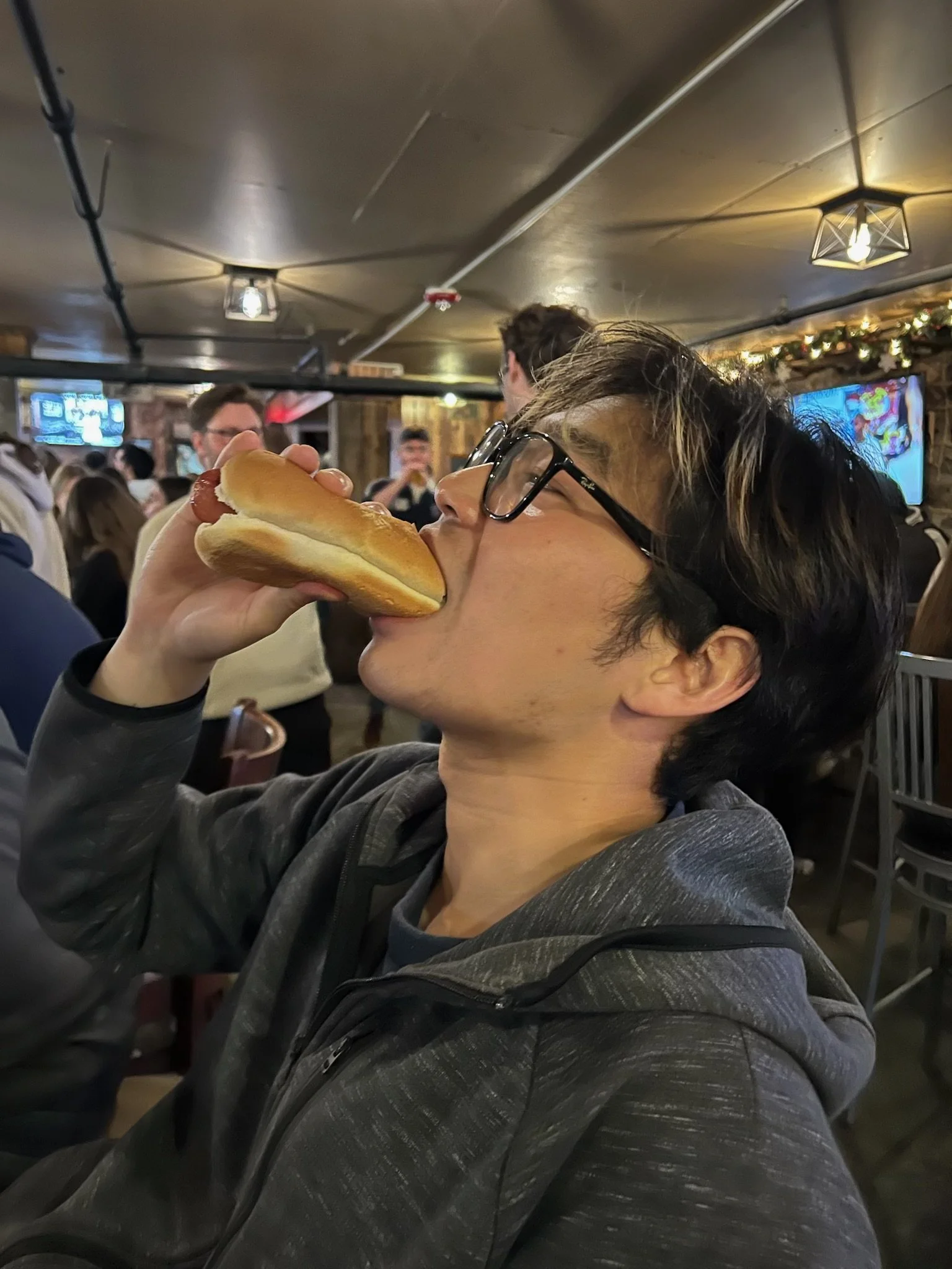 Person with glasses and dark hair wearing a gray hoodie, holding and eating a hot dog in a crowded indoor restaurant or bar.