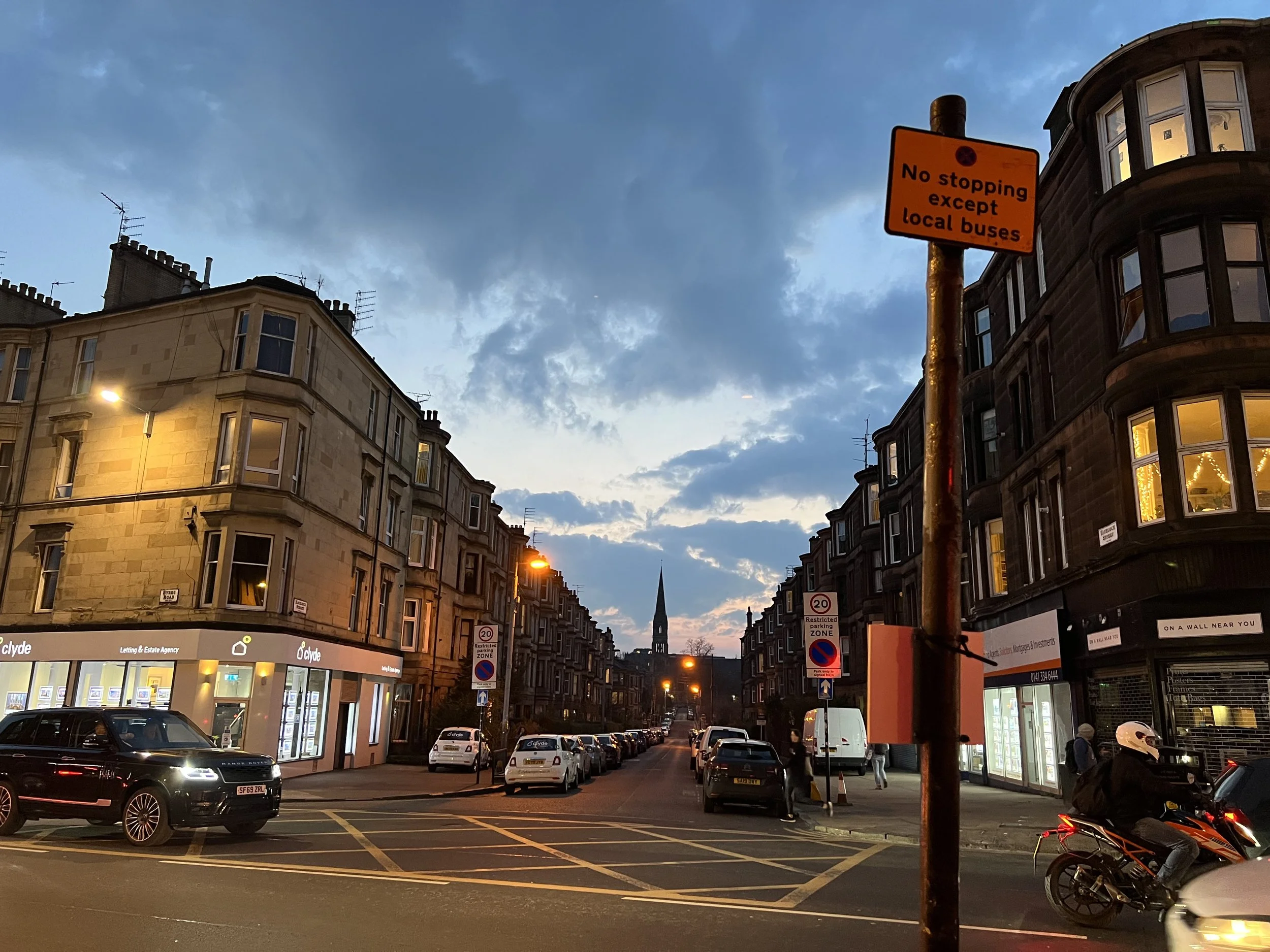 City street corner during twilight, with parked cars lining the street, buildings with illuminated windows, a street sign indicating no stopping except for local buses, and a church steeple in the distant background.