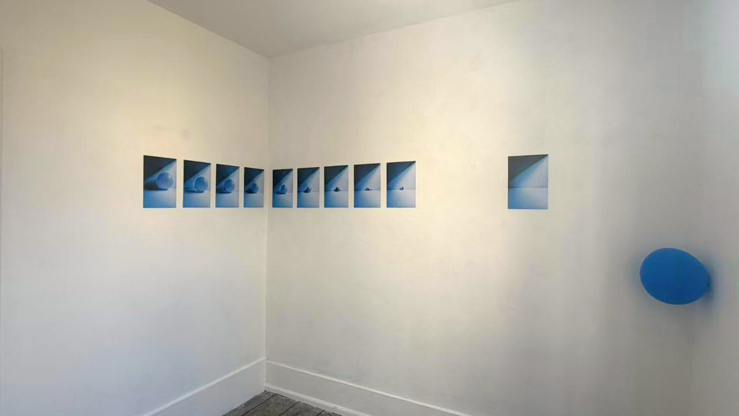 A series of nine rectangular photographs arranged in a horizontal row on a plain white wall. Each photo depicts a white spherical object with a shadow cast on a light blue surface, with the object gradually moving from the left to the right. There is