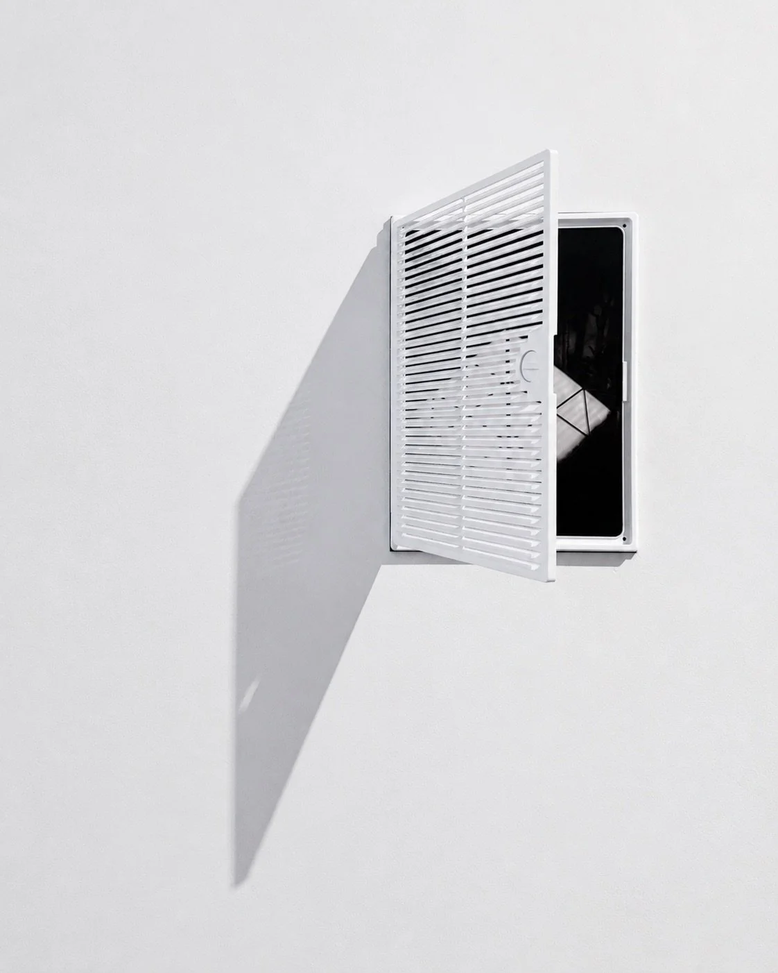 A white vent with a louvered cover on a white wall, casting a shadow.