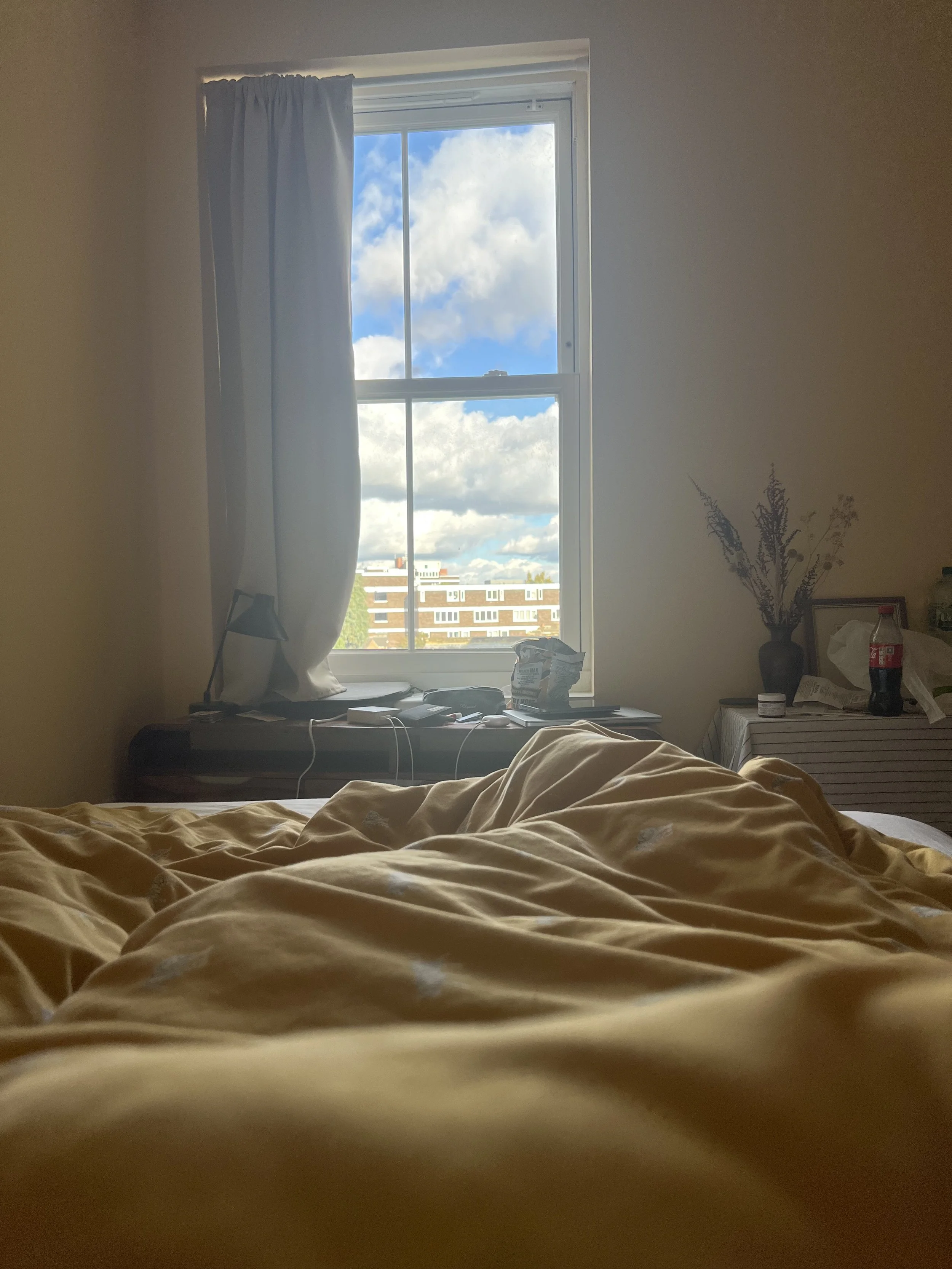 A bedroom with a window showing a cloudy sky and buildings outside. The bed in the foreground has yellow sheets, and there is a nightstand with a water bottle, snacks, and a vase with dried flowers on it.